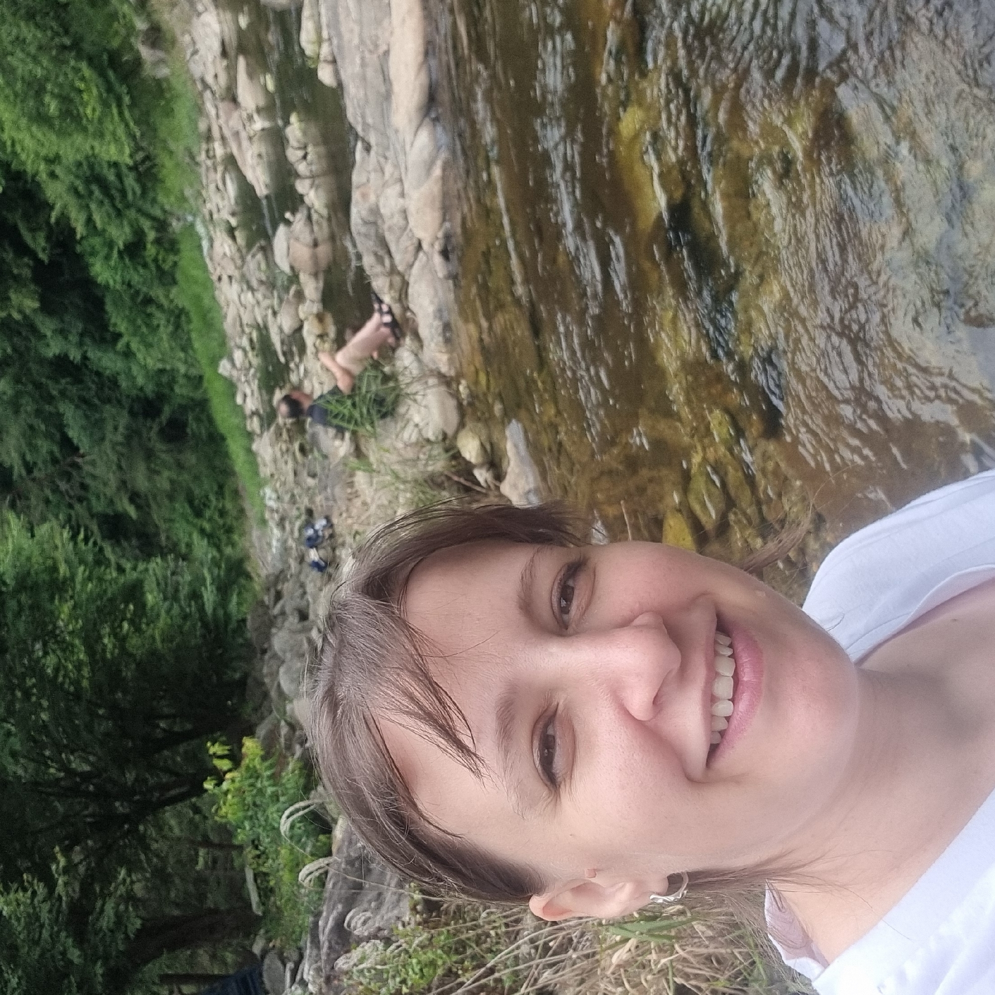 In an outdoor scene, a woman is pictured from the chest up, taking a selfie. She has fair skin, dark brown hair styled in a bob with bangs, and is wearing a white shirt and silver earrings. She is smiling broadly, showing her teeth. In the background, a shallow, rocky stream flows through a lush, wooded area. Two other individuals are visible in the background, sitting on the rocks by the stream. The environment is natural and appears to be a park or natural reserve. This image was captured in Mureungdowon-myeon, South Korea.