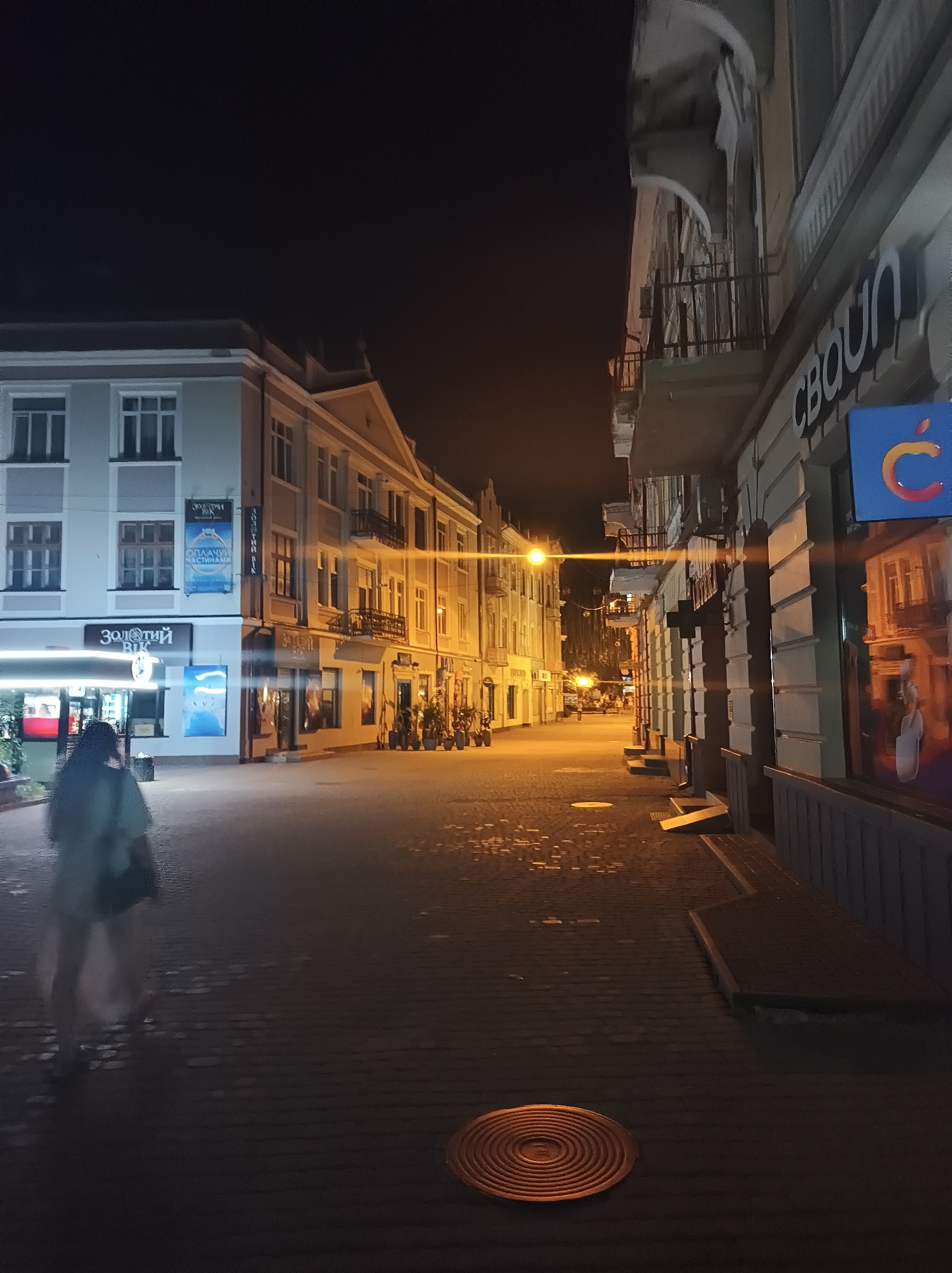 The image shows a street scene at night in Ternopil, Ukraine. The street is paved with cobblestones and is lined with buildings. On the left side of the street, there is a light-colored building with several windows. A banner for "Zolotyi Vik" (Golden Age) is displayed on this building, along with an advertisement for wedding rings. Below this, there is a shop with illuminated signs. A person wearing a light-colored jacket and dark pants walks away from the camera on the left side of the street. On the right side of the street, there is a building with architectural details, including balconies and arches. A sign with a stylized "C" logo is visible on this building. The street is illuminated by streetlights, casting a warm glow. There are some potted plants placed outside some of the shops on the right. A circular metal grate is visible on the cobblestone pavement in the foreground. The overall atmosphere is that of a quiet urban street at night.