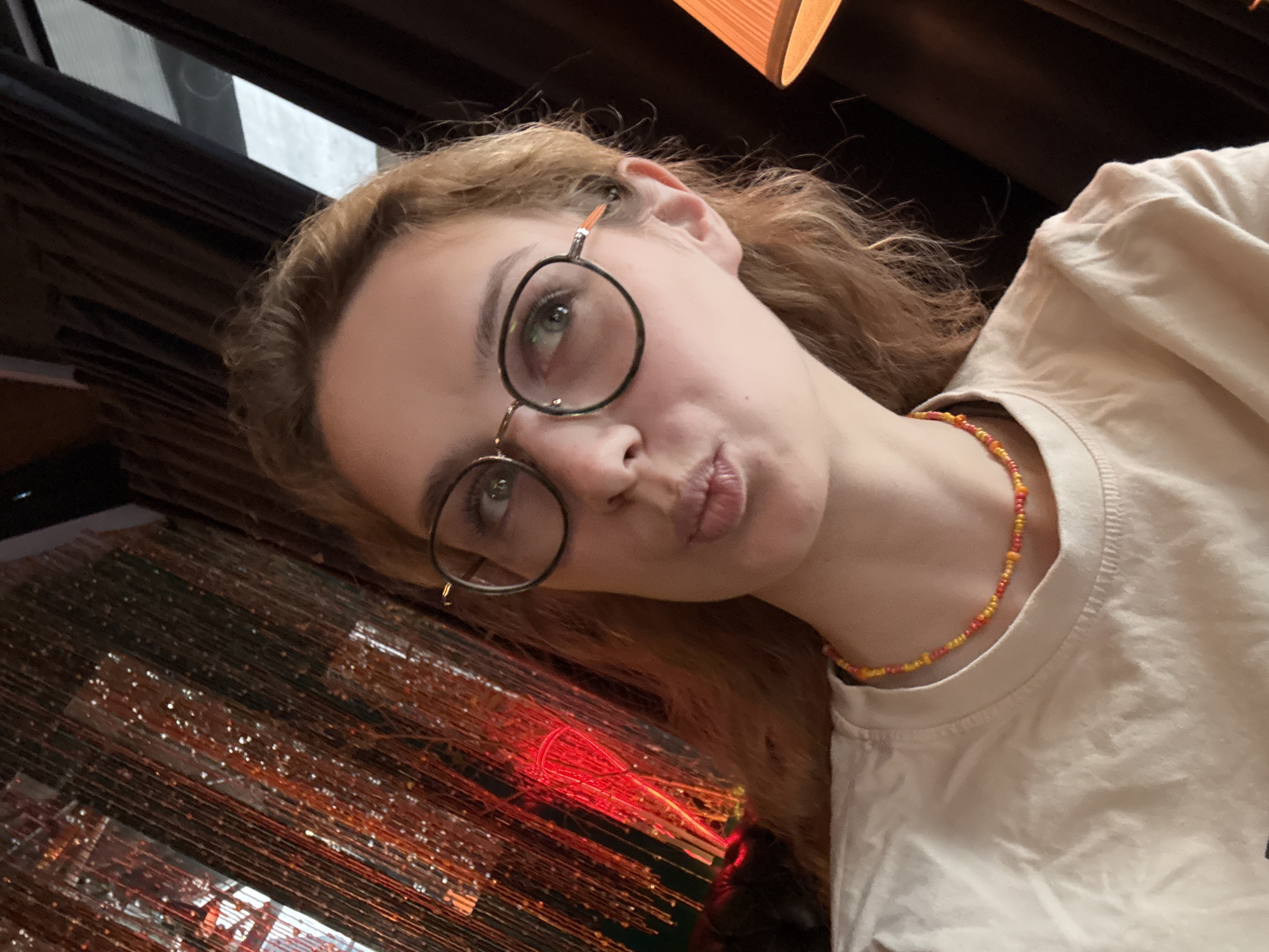 A selfie of a young woman wearing round glasses and a colorful beaded necklace. She is pursing her lips slightly. The background contains an indistinct decorative element made up of hanging strands that appear to be lit by colored lights. It's hard to identify a specific location in Belgrade without more information.