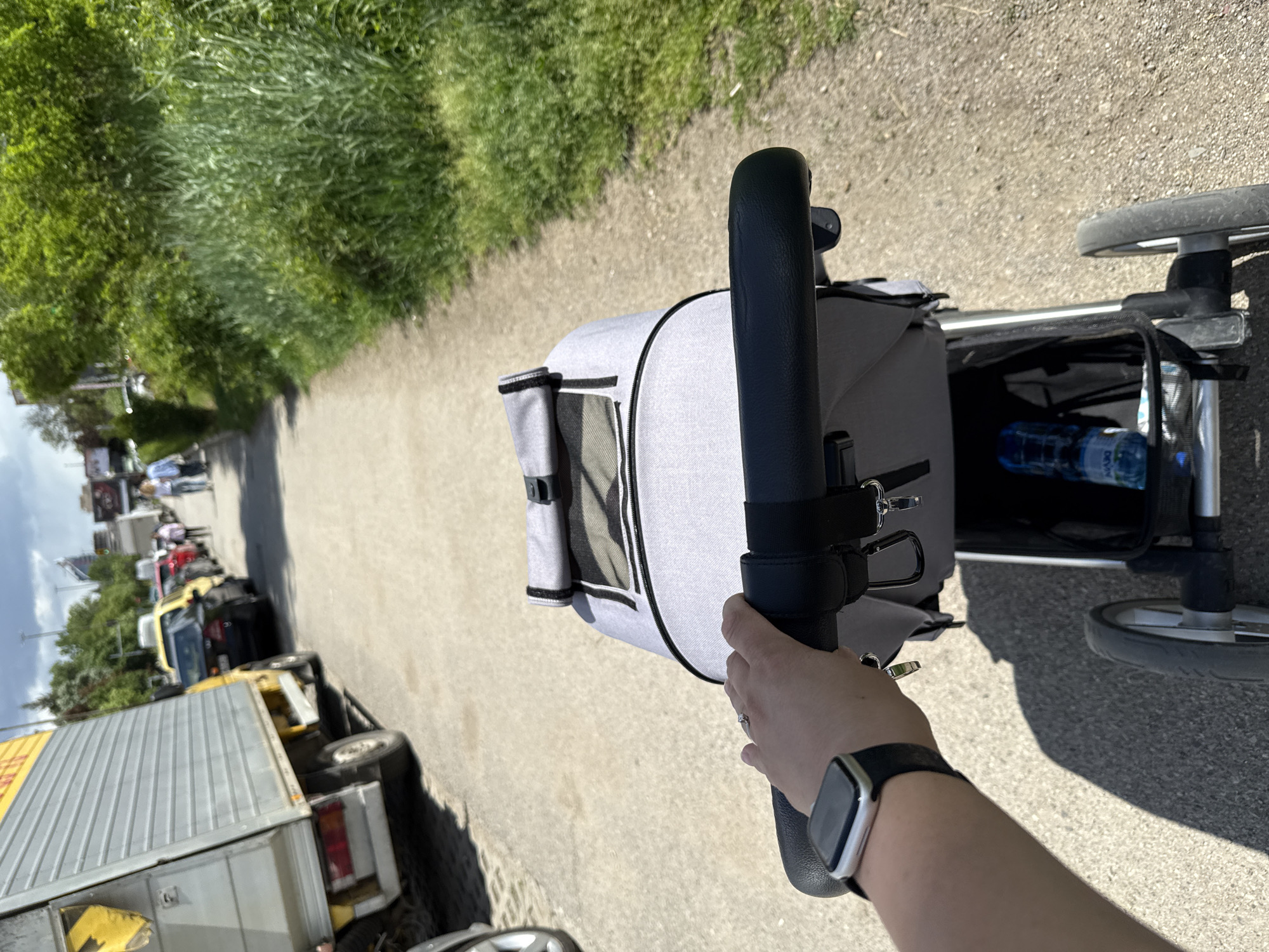 A person is holding the handle of a grey and black baby stroller, which appears to be on a paved path. To the left of the path is a line of vehicles, including a truck with a grey container and a yellow cab. There are also some people in the distance. The path is bordered by a grassy area with green foliage. In the stroller's storage area is a bottle of water.
The location is identified as Sofia, Bulgaria, based on available information.