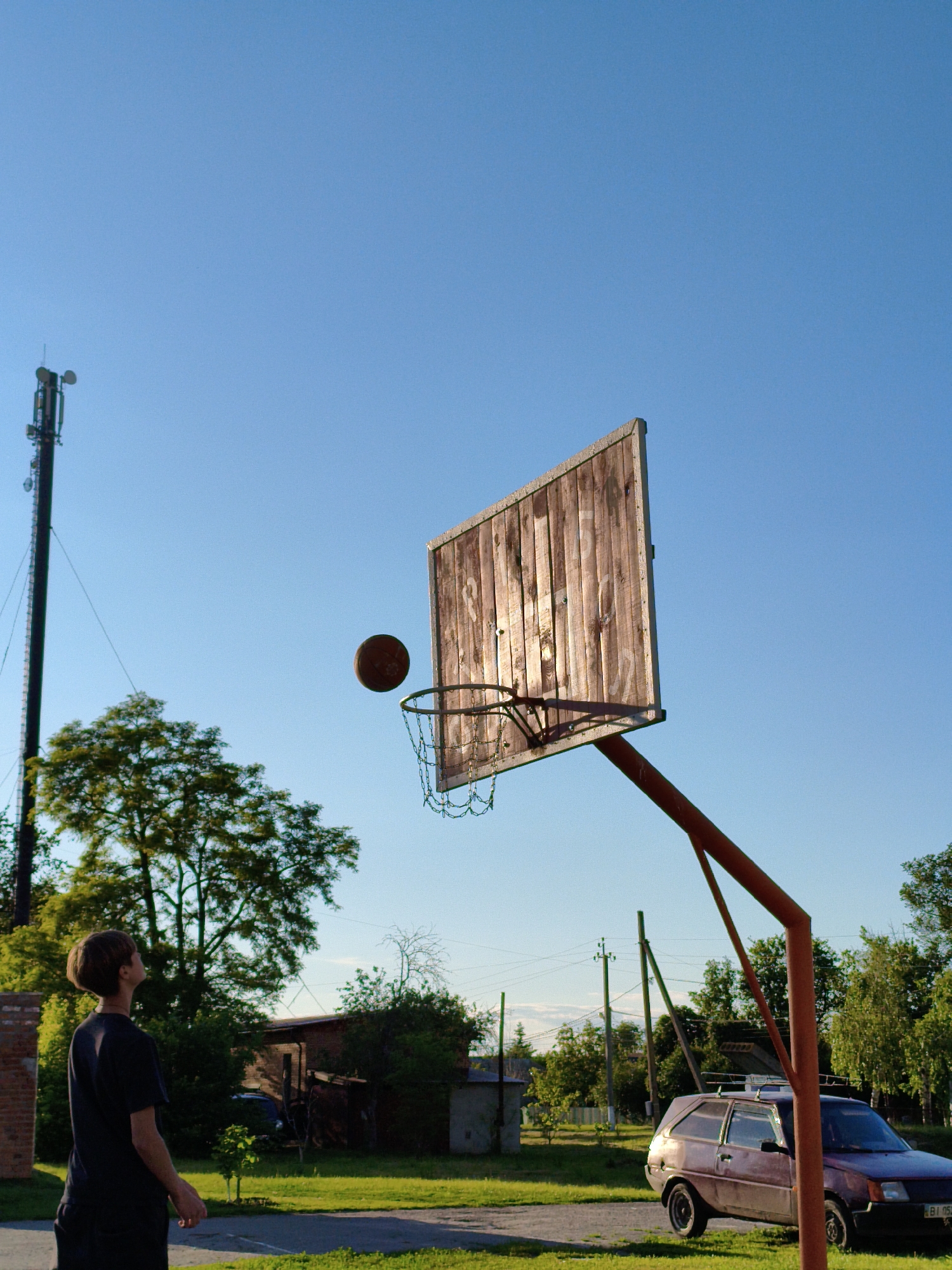 This outdoor photograph depicts a young person playing basketball in Ustyvytsia, Ukraine, during what appears to be the late afternoon or early evening, characterized by warm, low-angle sunlight. The sky is a clear, vibrant blue, occupying a significant portion of the upper frame.
The main subject is a person with short, dark hair, wearing a dark t-shirt and shorts, standing on a paved surface. They are seen from a side-back angle, looking upwards towards a basketball hoop. An orange-brown basketball is captured in mid-air, positioned just above the metal rim of the hoop, which features a chain net. The backboard of the hoop is constructed from weathered wooden planks and is brightly illuminated by the sun. The entire hoop assembly is supported by a reddish-orange metal pole.
The setting appears to be a casual, possibly makeshift, basketball court in a residential or semi-rural environment. In the background to the left, a tall cell phone tower stands prominently against the sky, with lush green trees and a brick building visible beneath it. To the right, an older, dark-red hatchback car is parked near the base of the basketball hoop's support pole. The car's license plate shows visible Cyrillic characters, specifically "BI 0524 BI". Additional trees, utility poles with power lines, and modest buildings or sheds are scattered throughout the background. The ground transitions from a paved court area to grass and dirt. The scene conveys a moment of focused recreational activity under pleasant, sunny weather.