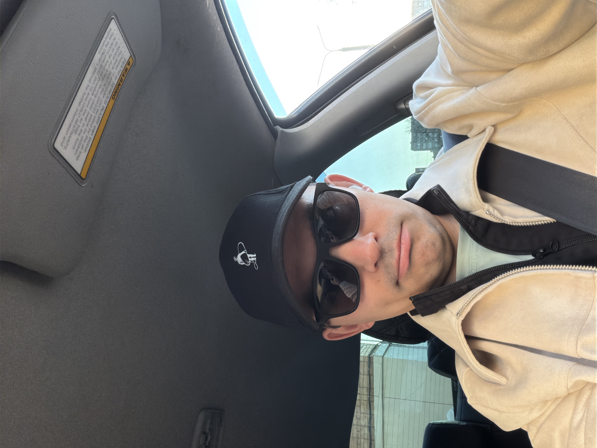 Here is a detailed description of the image:
A selfie taken from inside a car, likely in Izhevsk, Russia (based on possible metadata not visible). The main subject is a man wearing sunglasses and a black baseball cap with a white logo, possibly a caricature. He is also wearing a light-colored jacket or coat with a black zipper. The angle suggests he is either the driver or a passenger taking the photo towards the upper-left corner of the car's interior. The car's interior is primarily gray, with a visible sun visor and a warning label adhered to it. The exterior view through the car window is indistinct, but suggests a urban scene with buildings and daylight. The man appears to be looking directly at the camera.