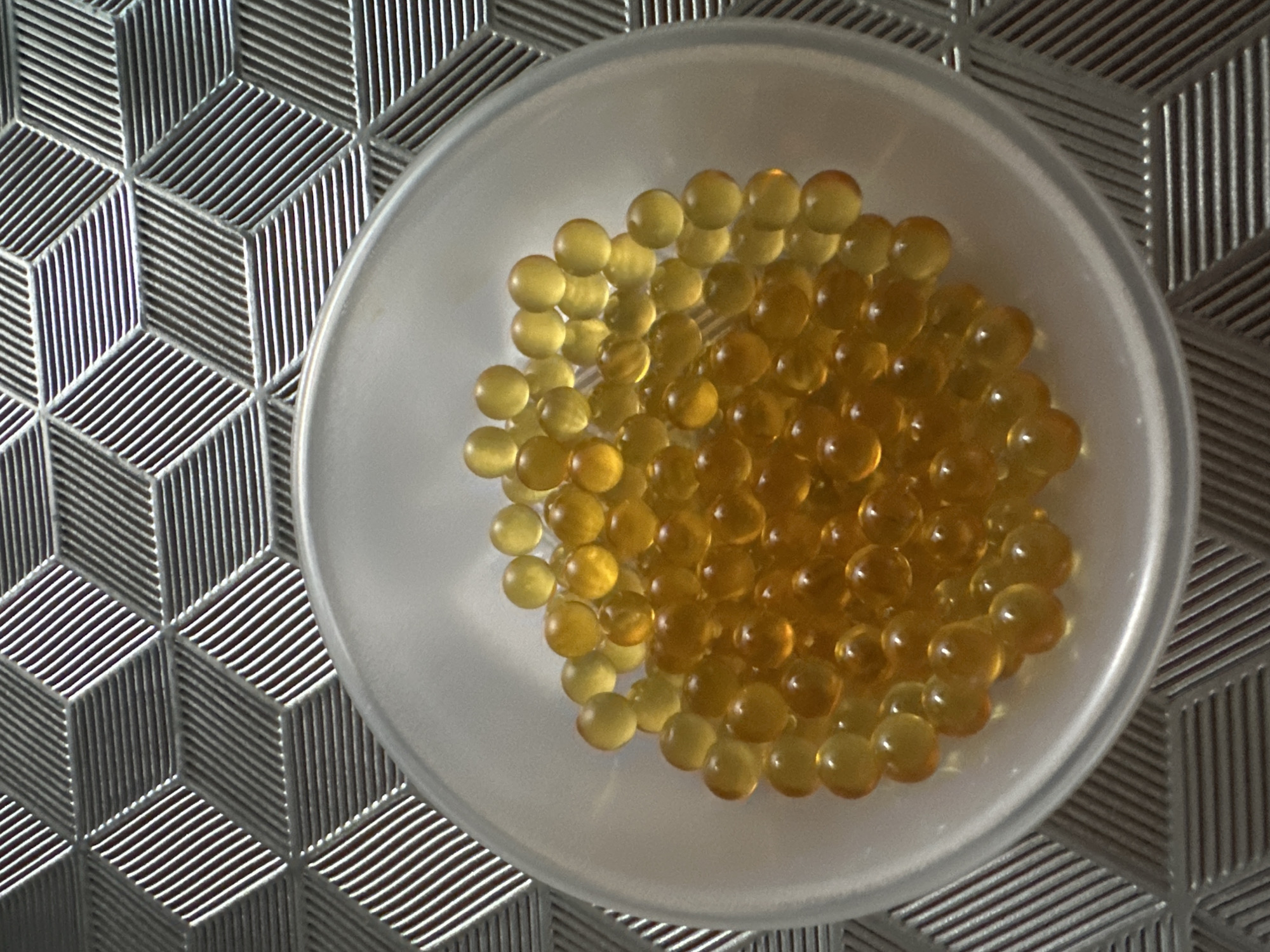 The image shows a translucent white bowl filled with numerous amber-colored, spherical gel capsules. The bowl is positioned on a surface with a geometric, cube-like pattern consisting of raised silver lines. The location is unspecified and not identifiable as Oryol, Russia from the visual information provided.