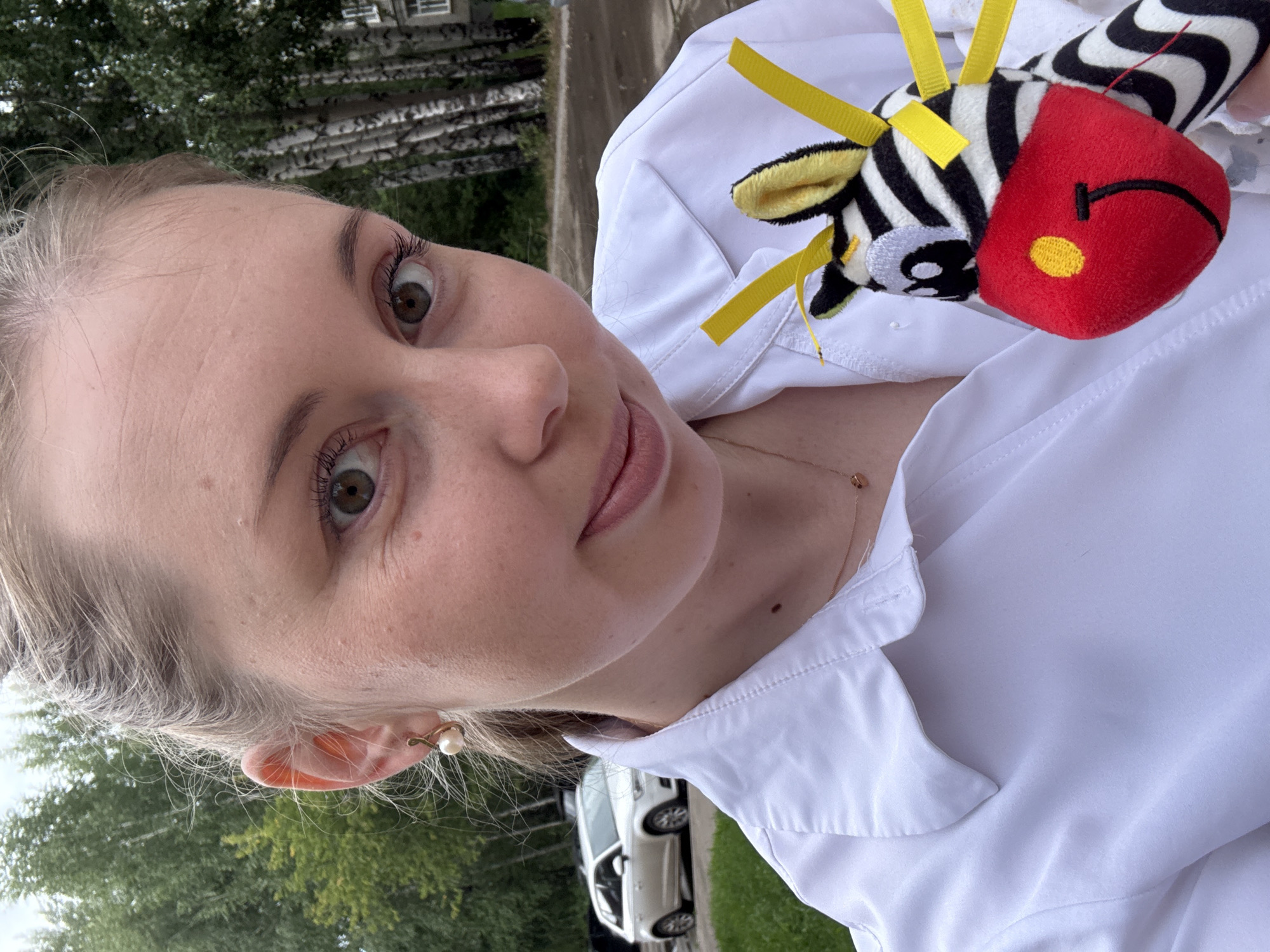 In this medium shot, a young woman with blonde hair pulled back and wearing a white collared shirt smiles directly at the camera. She has a pearl earring in her left ear and a delicate rose-gold necklace with a heart-shaped pendant. The woman holds a stuffed zebra toy with a red muzzle, yellow ears, and black and white stripes. Yellow ribbons are attached to the zebra's head. In the background, trees and a white car are visible, suggesting an outdoor urban setting. The lighting is natural and the image appears to be taken during the daytime.