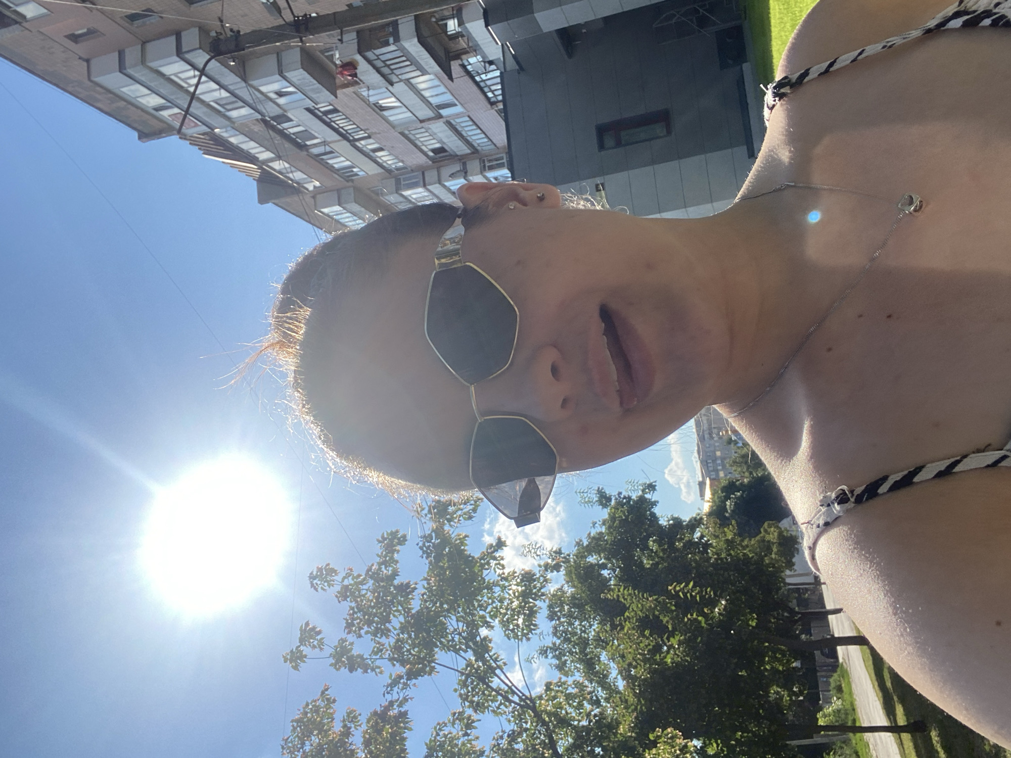 The media file is a selfie taken by a person outdoors on a sunny day. The subject is a young person wearing sunglasses and a halter top with a black and white pattern. They have a necklace with a small, geometric pendant. The background shows a clear blue sky, the sun shining brightly, and some trees. In the distance, there are buildings that appear to be apartment complexes. The image is captured from a low angle, looking up towards the subject and the sky. The overall impression is one of a casual, outdoor moment, possibly during warm weather given the clothing and bright sun. The lighting suggests it is daytime, likely midday or early afternoon due to the intensity of the sunlight and the clear sky. There is no visible text in the image.