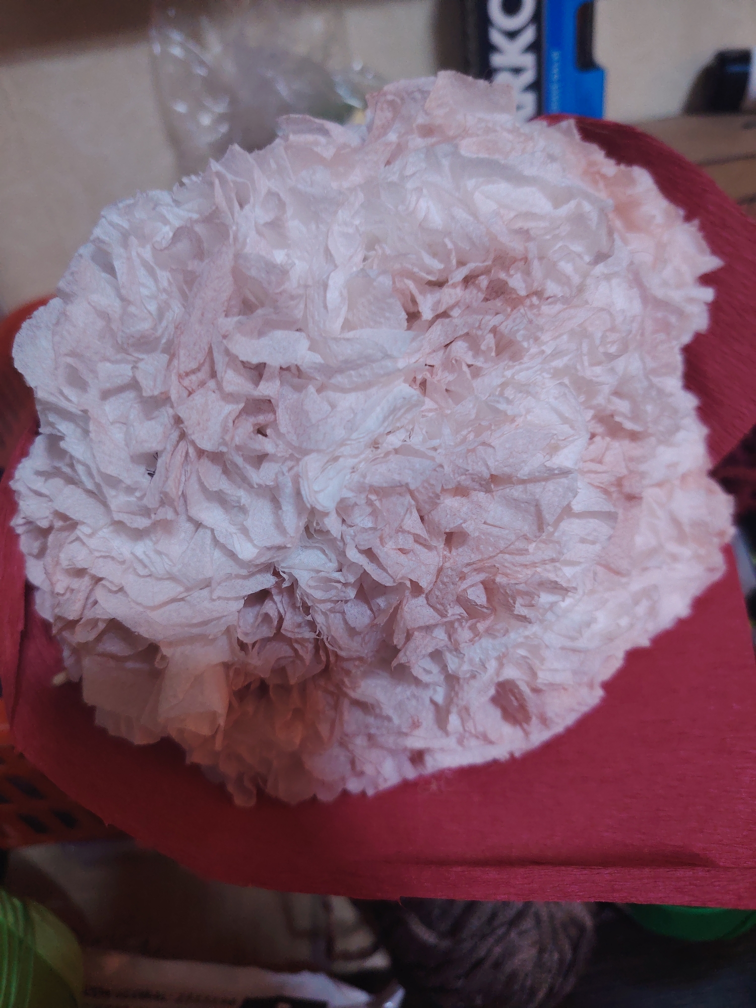 A close-up, overhead shot shows a light pink, crinkled paper pom-pom resting on a textured red surface. The pom-pom appears to be made of tissue paper, with individual layers of paper bunched and twisted to create a floral or fluffy texture. Some subtle reddish-pink discoloration is visible on the paper. The red surface underneath has a crepe-like texture and a deep, rich color. In the background, out of focus, is a plastic-wrapped item and a blue object with white lettering, possibly a container or packaging. The overall setting appears to be indoors, with soft lighting.