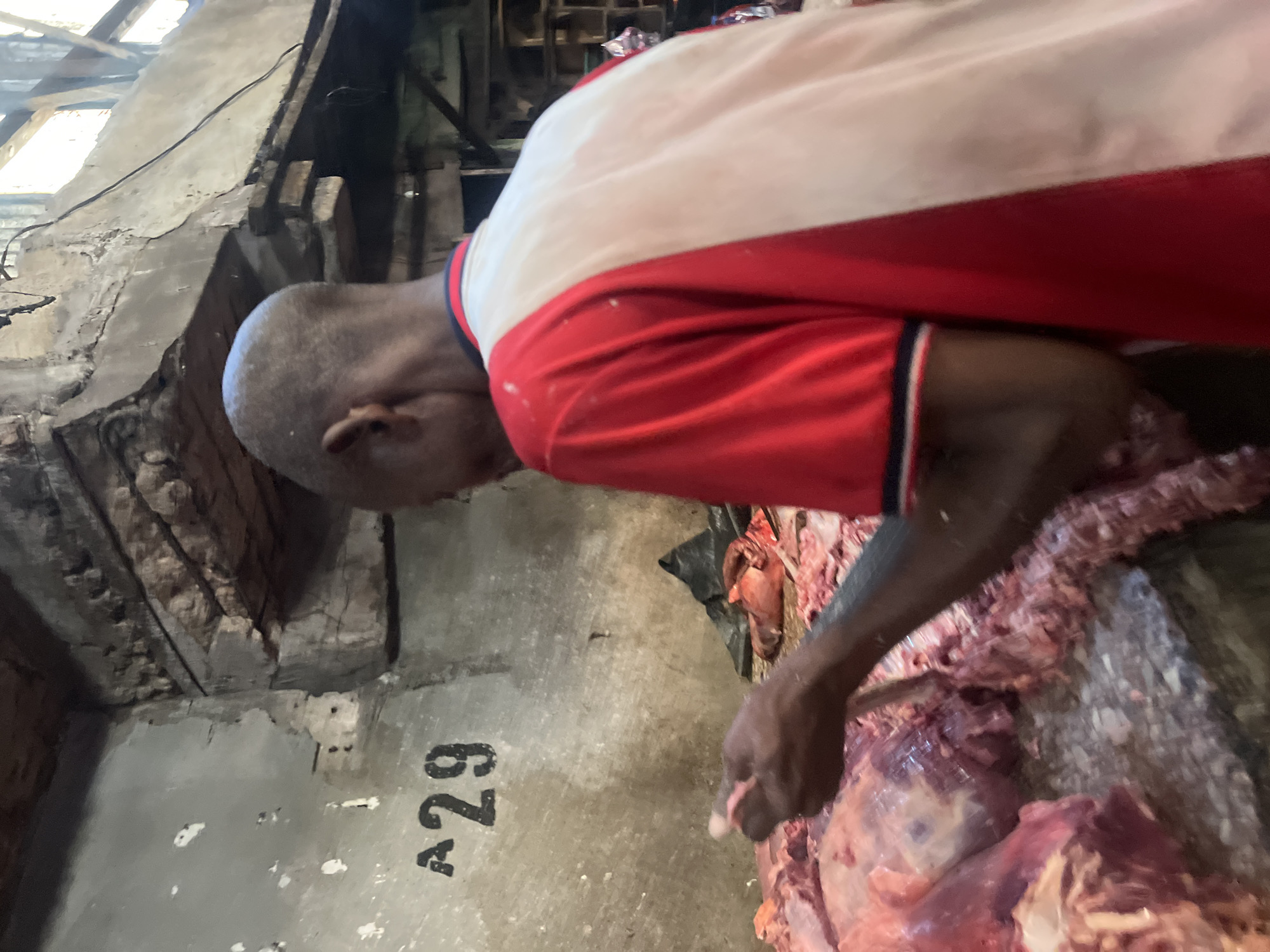 A man with a shaved head, wearing a red and white shirt, is cutting meat. The meat is situated on a gray surface marked with the number "A29." The setting appears to be an enclosed space, possibly a butcher shop, in Nkpor, Nigeria, given the visual information available. The background includes structural elements of the building, suggesting an older, possibly dilapidated, interior. The focus is on the act of cutting meat, with details of the man's actions and the surrounding environment visible.