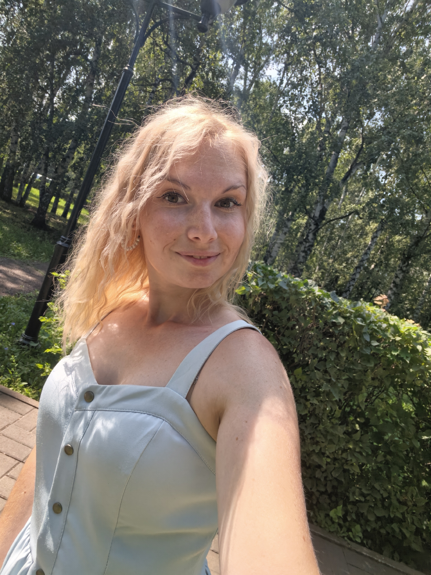 A fair-skinned woman with blonde, wavy hair and light brown eyes is pictured from the chest up, outdoors on a sunny day. She is wearing a light blue, sleeveless dress with buttons down the front. She has freckles across her nose and cheeks, and is wearing pearl earrings. She is smiling at the camera. Behind her is a park-like setting with green trees and foliage, and a lamppost is visible to her left. The ground in front of her appears to be paved with brick. The location is identified as Sterlitamak, Russia.