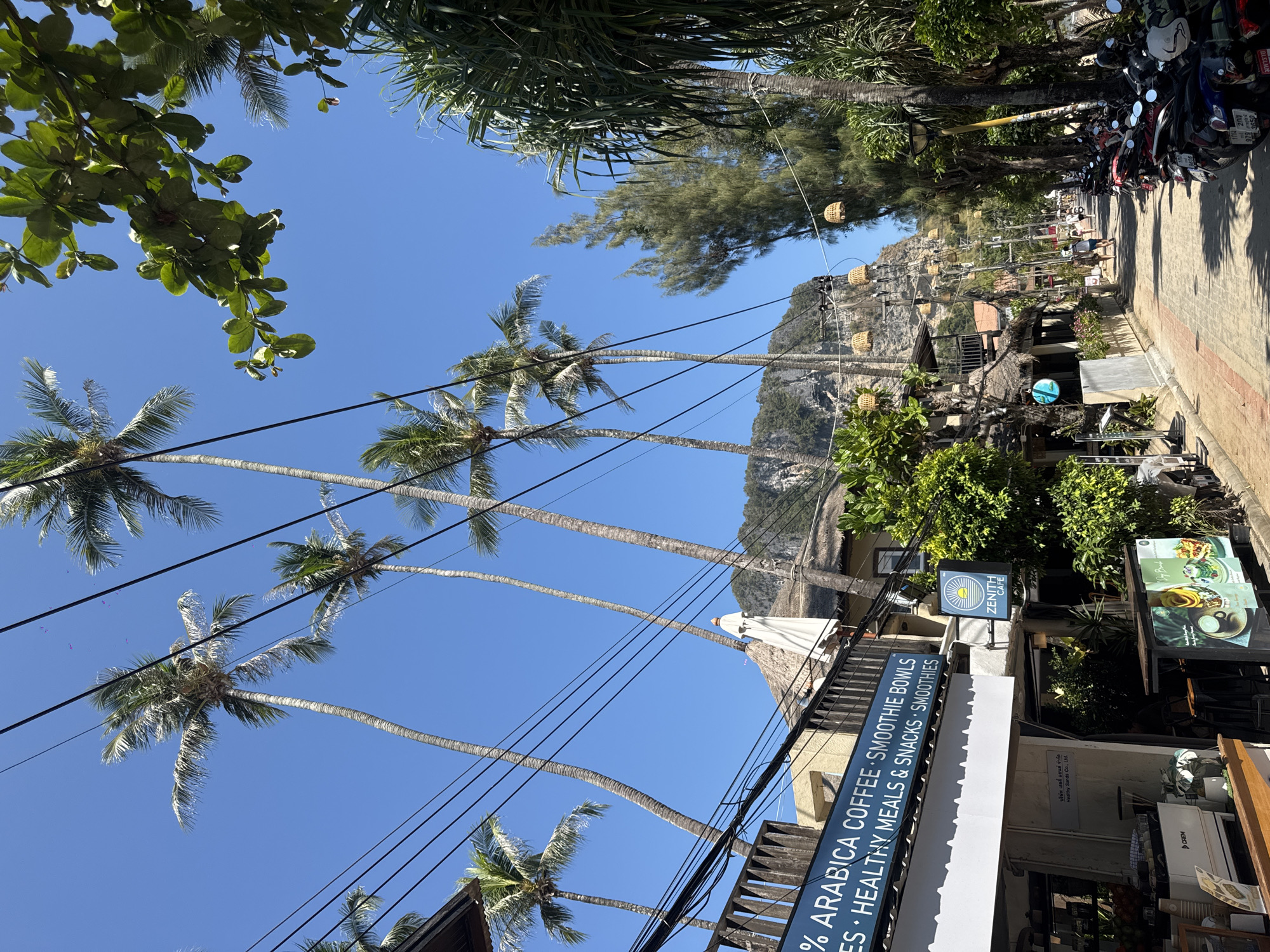 The image shows a street scene in Ban Ao Nang, Thailand. Tall palm trees with long, slender trunks reach into a bright blue sky, with power lines running between them. On the right side of the street, several parked motorbikes line the paved road. A building with signage reading "% ARABICA COFFEE • SMOOTHIE BOWLS • HEALTHY MEALS & SNACKS • SMOOTHIES" is visible, along with a sign for "ZENITH CAFE". A small restaurant or cafe area is set up, featuring menus and plants. In the background, a cliff or rocky hill is visible. Lanterns hang from the power lines further down the street, and pedestrians can be seen walking along the sidewalk. Lush green foliage is present in the foreground and background.