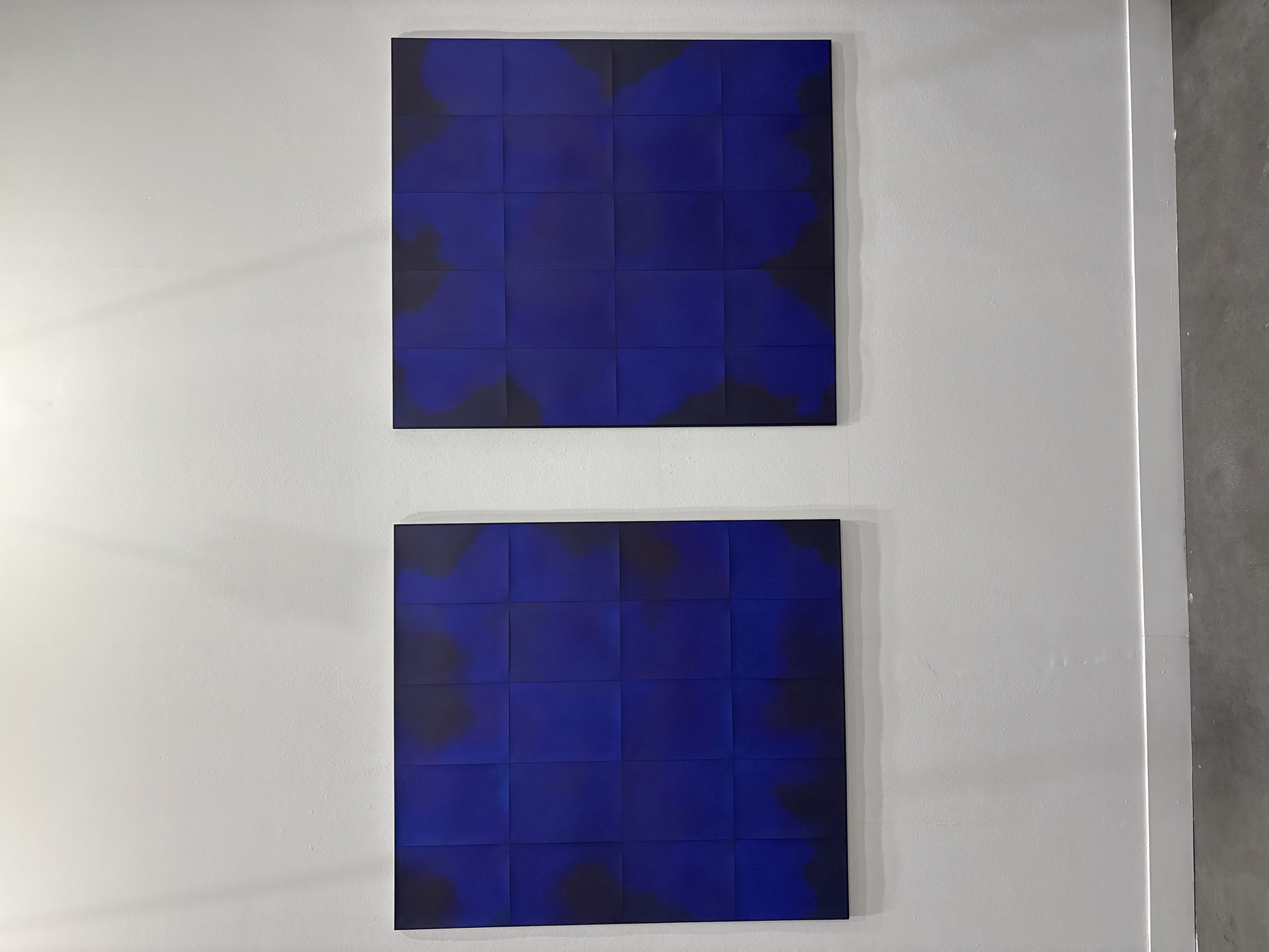 The image depicts an interior scene, likely an art gallery or exhibition space in Milan, Italy, showcasing two identical abstract artworks mounted vertically on a pristine white wall. Each artwork is a large square, composed of a 4x4 grid of smaller square panels. The pieces feature a deep, rich blue color interspersed with amorphous, cloud-like shapes of black, which create a sense of depth and movement across the tiled surfaces. These black areas appear as darker tones or shadows within the blue field. Both artworks are framed with a slender black border and are positioned one above the other, separated by ample white wall space.
The white wall exhibits subtle textures and is softly lit from the left, casting faint shadows that highlight its surface. To the far right, a narrow vertical white strip, possibly part of a door frame or architectural trim, separates the main wall from an adjacent gray surface and a concrete-like floor. No people are visible, and there is no discernible text within the image. The setting conveys a minimalist and contemplative atmosphere, focusing solely on the visual impact of the modern artworks.