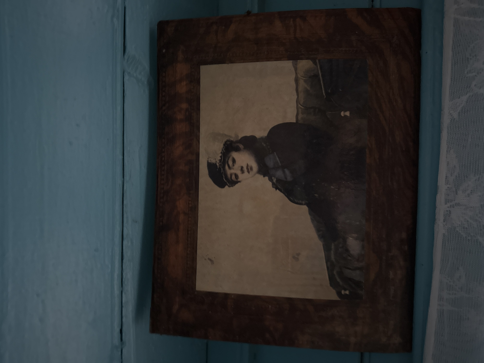 A framed sepia-toned portrait of a young woman in historical attire is displayed on a light blue wooden surface, possibly a shelf or ledge, in Sosnovskoye, Russia. The woman, positioned in the center of the frame, is depicted from the waist up, seated and looking towards the viewer. She wears a dark jacket or dress with a distinctive hat featuring a dark brim and a lighter-colored, fluffy crown. A blue ribbon or bow is visible at her neck. The background is indistinct, appearing as a muted interior. The frame of the portrait is made of dark wood with an embossed pattern along its edges. Below the framed portrait, a white lace curtain is visible, obscuring the lower portion of the scene. The light blue surface is textured, suggesting painted wood.