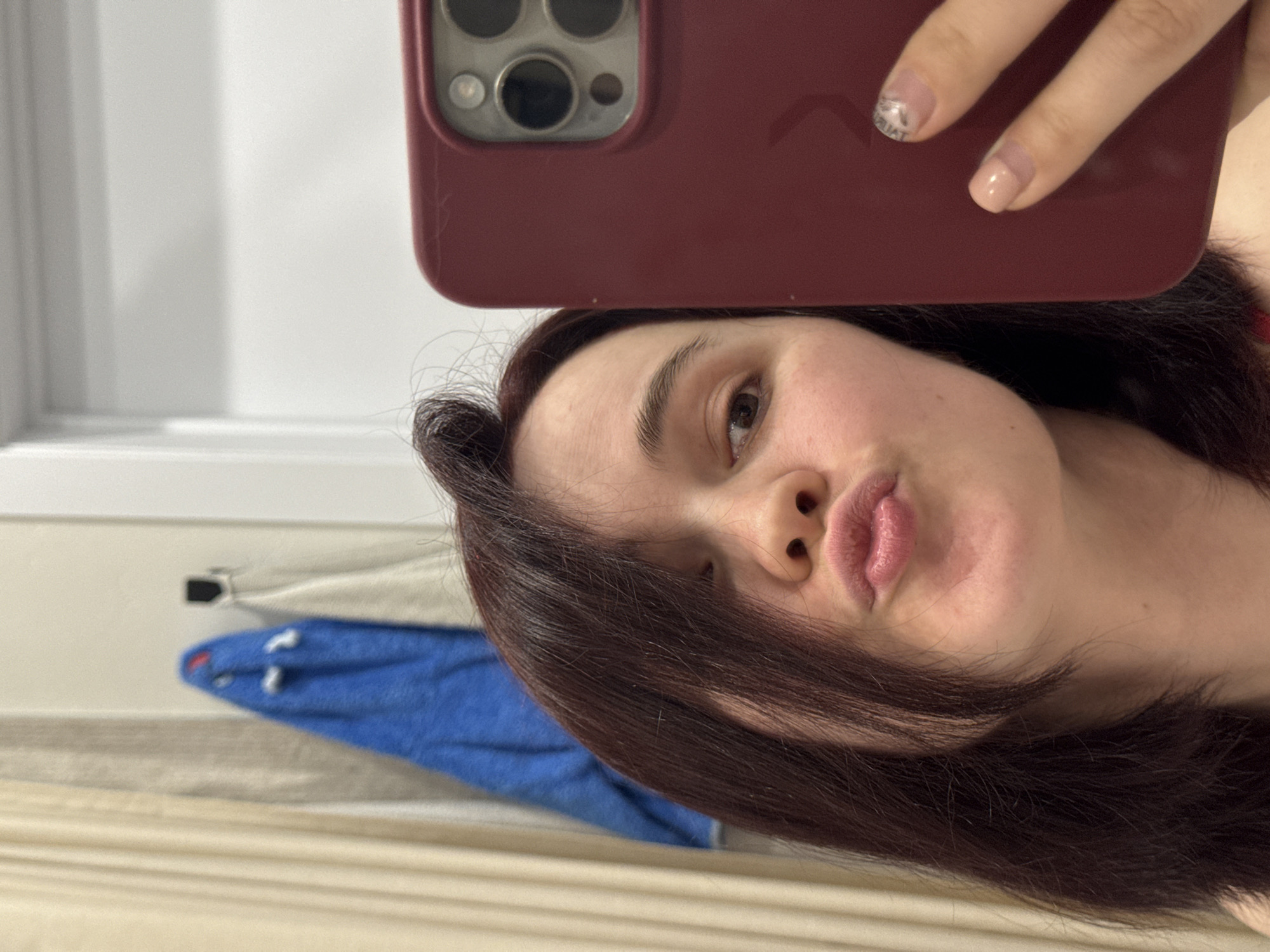 A selfie captures a young woman indoors, holding a dark red smartphone case with her right hand. The image is rotated 90 degrees counter-clockwise, showing the woman's head tilted. She has dark, medium-length hair that partially covers her forehead and cheeks. Her skin appears natural and unblemished, and she has brown eyes and dark eyebrows. She is making a playful "duck face" with her lips slightly puckered.
Her right hand, holding the phone, has manicured nails. One nail shows a design that appears to read "TRUTH" or a similar word, reversed, while another features a French tip style. The camera array of the smartphone, featuring multiple lenses, is visible at the top of the frame.
The background consists of a bright, light-colored wall or window frame on the left, suggesting daylight illumination. Below, a lighter, textured surface, possibly bedding or a curtain, runs horizontally across the bottom of the frame. A blurred blue object, which resembles a folded blanket or towel with white details, is visible in the lower-left background. The overall scene conveys a casual, personal moment captured by a self-portrait. No specific cues within the image indicate the provided location of Izhevsk, Russia.