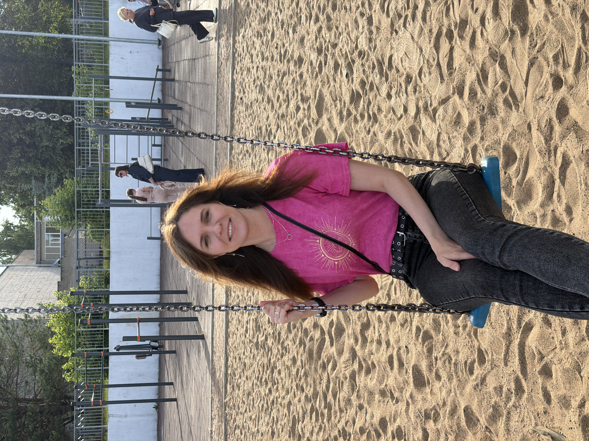 A young woman with long, brown hair is sitting on a swing with a blue seat, holding onto the metal chains. She is wearing a pink t-shirt with a sun-like graphic and black jeans with a black belt. She is smiling at the camera. The swing is located in a sandy playground. In the background, there are some exercise bars and a fence. Further in the background, there are buildings and trees. The lighting suggests it is daytime, possibly late afternoon, with soft shadows. There are other people visible in the background, walking near the playground. The overall mood appears relaxed and happy.