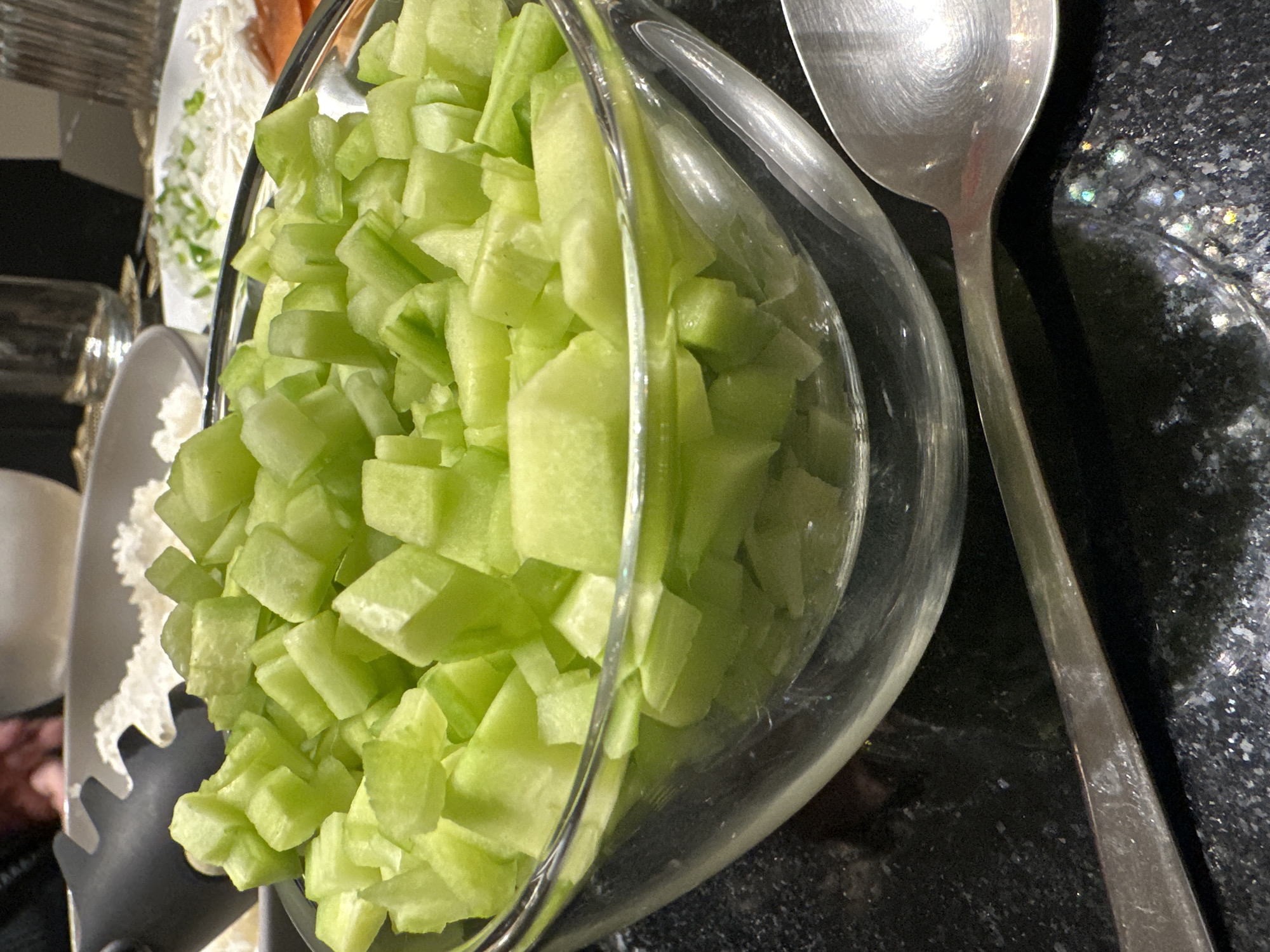 A close-up shot shows a clear glass bowl filled with small, uniformly cut cubes of a pale green fruit, likely honeydew melon or a similar type. A silver spoon rests on the dark, possibly granite, surface to the right of the bowl. To the left, out of focus, are other dishes containing what appears to be shredded cheese or vegetables. The setting seems to be a kitchen counter or tabletop. The location in Mexico City, Mexico, cannot be verified from the image.