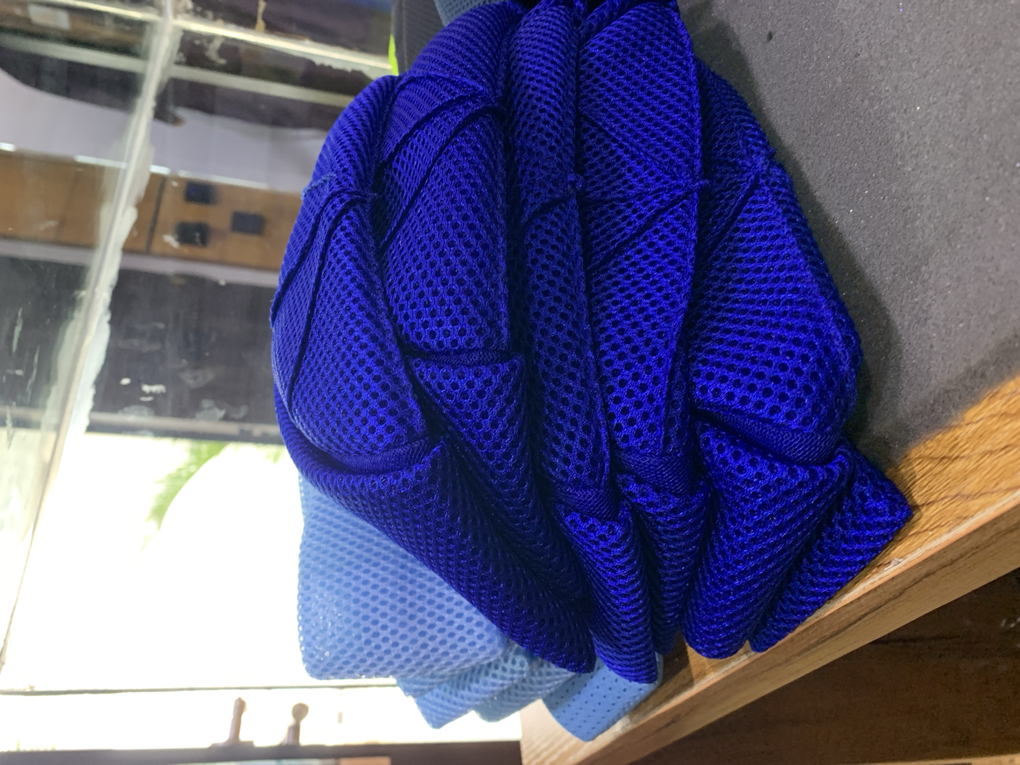 In Gura Top, Nigeria, a stack of folded blue mesh fabric is displayed on a wooden shelf. The fabric is layered, with the topmost pieces a deep royal blue, and the bottom layers a lighter shade of blue. The mesh pattern is visible throughout, creating a textured surface. The background appears to be a room with reflective surfaces, possibly mirrors or glass, showing reflections of the environment. The lighting suggests an indoor setting, possibly a shop or storage area, with natural light filtering in from the left.