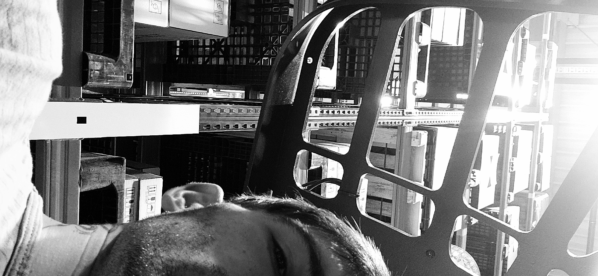 The black and white image depicts an industrial scene inside what appears to be a warehouse or distribution center in Plovdiv, Bulgaria. The perspective is from a low angle, possibly from within a forklift or similar high-reach machinery, looking upwards towards rows of industrial shelving.
In the foreground, the top of a person's head, including their ear and part of their face with stubble and an eye, is visible on the lower left. A large, dark metallic structure with geometric cutouts dominates the right side of the mid-ground, likely part of the machine from which the photo is taken, acting as a protective cage or frame.
The background features an extensive industrial racking system, with multiple levels holding various cardboard boxes. The shelves themselves are constructed from metal, showing repetitive patterns of triangular cutouts. Several boxes display shipping symbols, including "this side up" arrows and a fragile (glass) icon. One box in the upper left also shows symbols resembling a speaker and a circular warning sign. A small, distinct black square is visible on a white horizontal support beam.
The scene is characterized by strong contrasts and reflections due to bright lighting, with an intense light source visible on the far right, creating significant glare. The overall impression is one of an active, well-stocked industrial storage environment. The time of day is not discernible due to the indoor setting and artificial lighting.