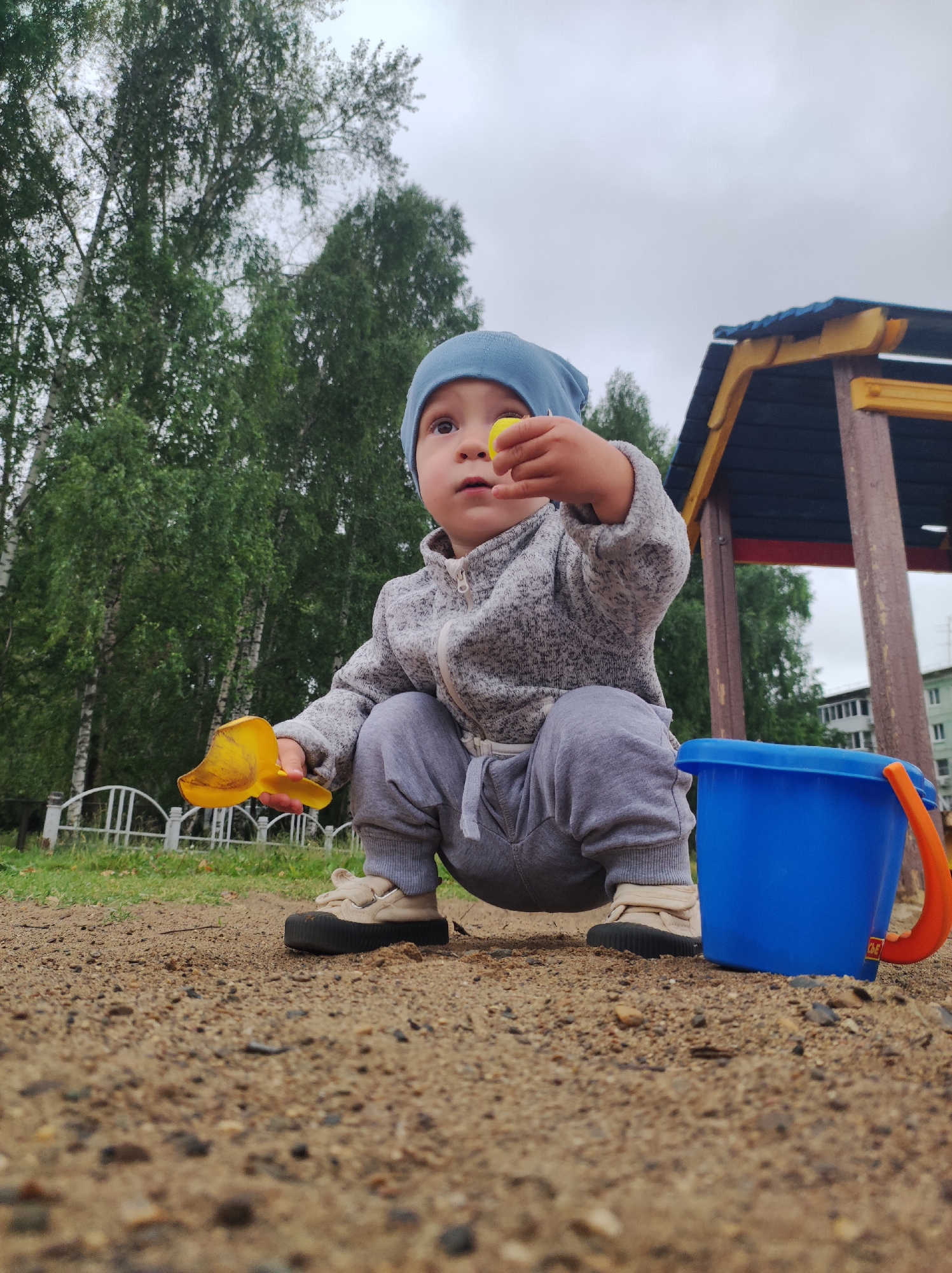A young child, likely a toddler, is crouching in a sandy playground area. The child is wearing a light blue hat, a grey hooded jacket, and grey pants. They are holding a yellow toy shovel in their left hand and a small yellow object in their right hand. Beside them, to the right, is a bright blue plastic bucket with an orange handle. In the background, there are trees with green foliage, a white fence, and part of a wooden play structure. The sky is overcast. The ground is covered in sand and small pebbles. This scene appears to be in a city park or residential courtyard, consistent with a public space in Chaikovsky, Russia.