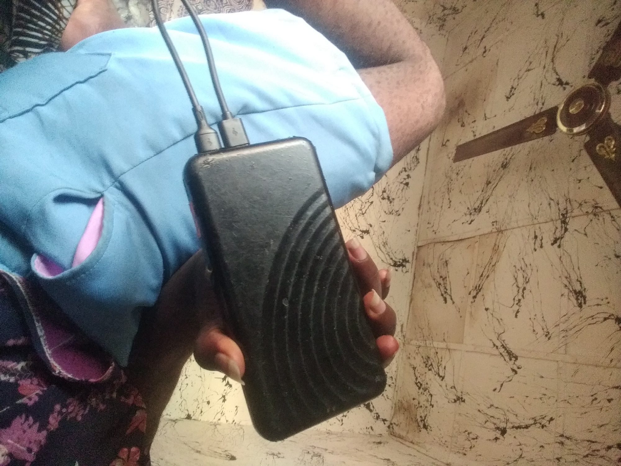The image depicts a close-up view of a person's legs and hand holding a black power bank. The person is wearing light blue shorts and a dark floral patterned garment. Two USB cables are connected to the power bank, one of which extends towards the person's lap. The background shows a textured ceiling with a brown ceiling fan. The scene is likely indoors, within a residential setting in Jalingo, Nigeria. The lighting appears to be natural or from an overhead source, casting soft shadows. The overall impression is one of everyday life, with a focus on personal electronics and clothing.