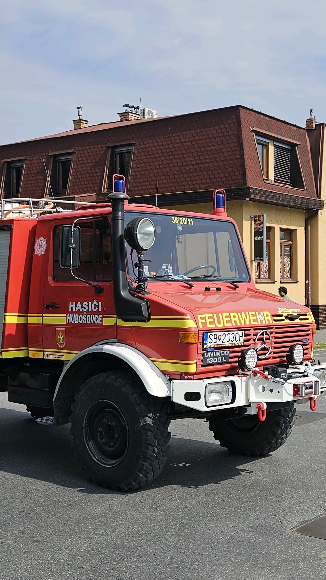 A red fire truck with yellow stripes and the words "HASIČI, HUBOŠOVCE" and "FEUERWEHR" is parked on a street in front of a building with a reddish-brown shingled roof. The fire truck has a large snorkel on the driver's side, a spotlight, and two blue flashing lights on the roof. The license plate reads "SB 203CH". The building appears to be a commercial establishment, possibly a shop or restaurant, with windows displaying signage. A person is visible in the background near the building entrance. The sky is partly cloudy. The image is taken from a low angle, focusing on the front and side of the fire truck.