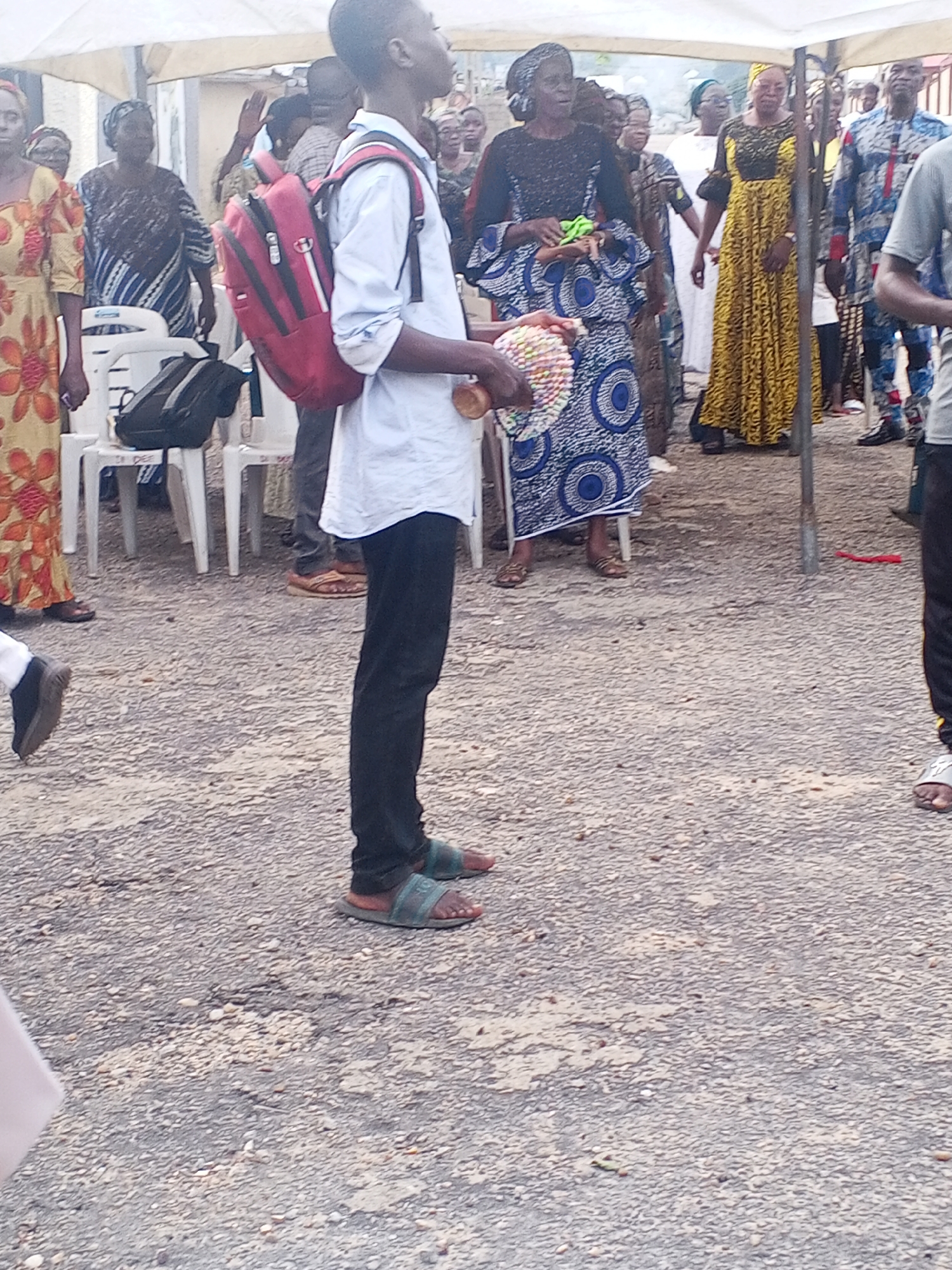 In Lokoja, Nigeria, a group of people are gathered outdoors under a white tent. A young boy in the foreground, wearing a light blue shirt, dark pants, and sandals, has a red backpack on and is holding a colorful object. He is facing to the right, looking slightly upward. Behind him, various individuals are dressed in traditional African attire. To his left, a woman in a yellow and orange patterned dress stands, and another woman in a blue and white patterned outfit is also visible. Several white plastic chairs are arranged in the background. The ground is covered with gravel and dirt. The atmosphere appears to be that of a community gathering or event.