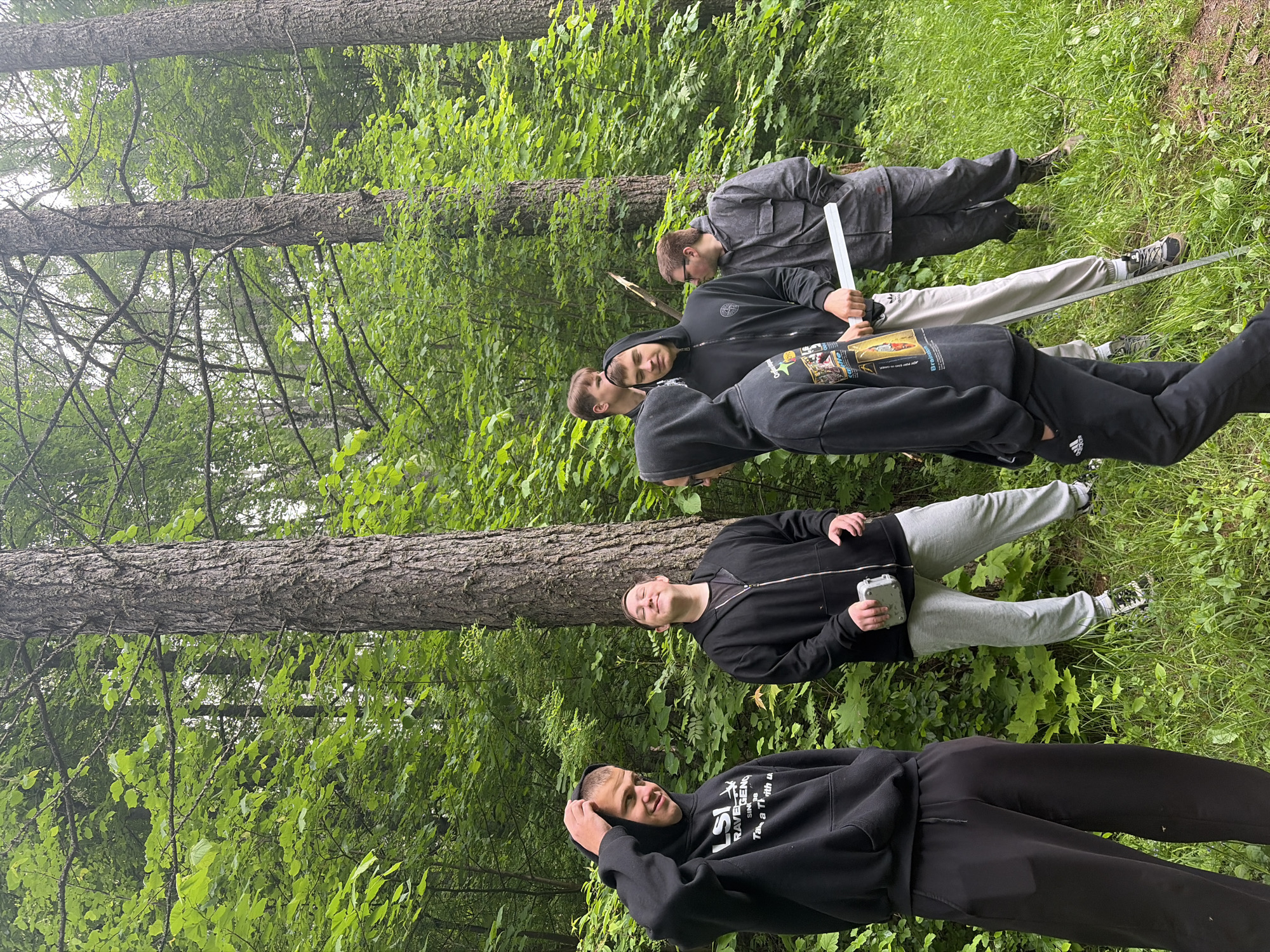 Six young men are standing in a lush green forest, likely participating in an outdoor activity or gathering. The scene is set in Красные Баки, Russia, during what appears to be daytime, possibly under an overcast sky, given the soft lighting. Tall coniferous trees with rough bark dominate the background and stand vertically, alongside deciduous trees with vibrant green foliage and dense undergrowth covering the forest floor.
The men are dressed in casual, athletic attire, primarily black hoodies and sweatpants, along with sneakers. In the foreground, slightly to the left, a young man with short hair smiles towards the camera, his hand resting on his head. He wears a black hoodie with "LSI RAVENCLAW" and smaller text like "Since 98" and "Take a Trip With Us" visible on it, paired with black Adidas sweatpants.
Behind him and slightly to his right, another young man looks upward, holding a white container or bottle in his hand. He is wearing a black zippered hoodie and light grey Adidas sweatpants. Further into the group, a man with a hood up has a serious expression, and next to him, another wears a black hoodie with a visible Stone Island patch on the sleeve, holding a long, white, rectangular object that resembles a prop or mock weapon. Another individual further back holds a wooden stick, while the furthest man in a dark grey jacket also holds a similar white rectangular object.
The men's expressions vary, from smiling and relaxed to more focused or contemplative, suggesting they are engaged in an activity that involves props, possibly a game, role-play, or team exercise. The overall atmosphere is one of a group enjoying the outdoors in a natural, wooded environment.