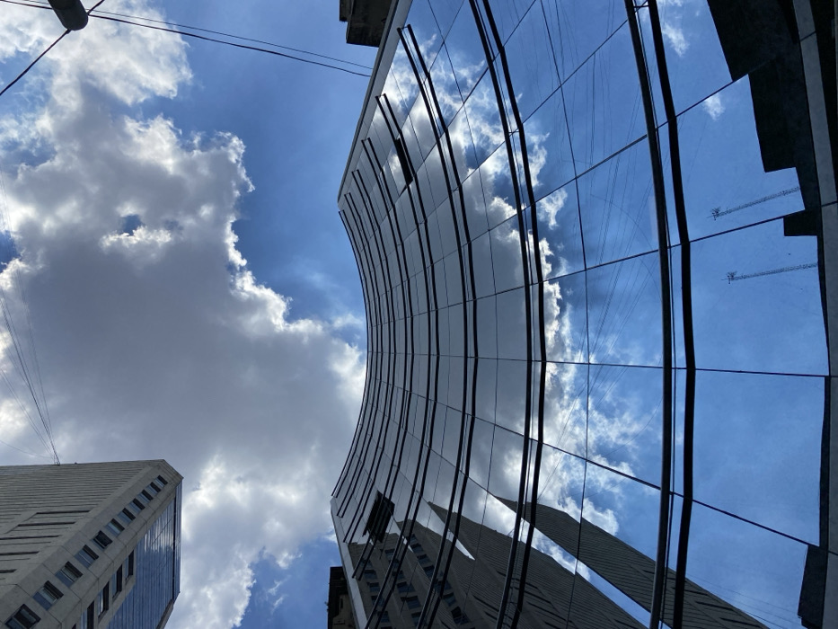 A low-angle perspective captures a modern glass skyscraper curving toward the sky. The reflective glass panels mirror the blue sky and scattered white clouds, creating a distorted reflection of the adjacent building. Power lines stretch across the upper portion of the frame. Another, more traditionally designed skyscraper with a grey facade and regular window pattern is visible in the lower left. The composition blends natural and urban elements, showcasing contemporary architecture.