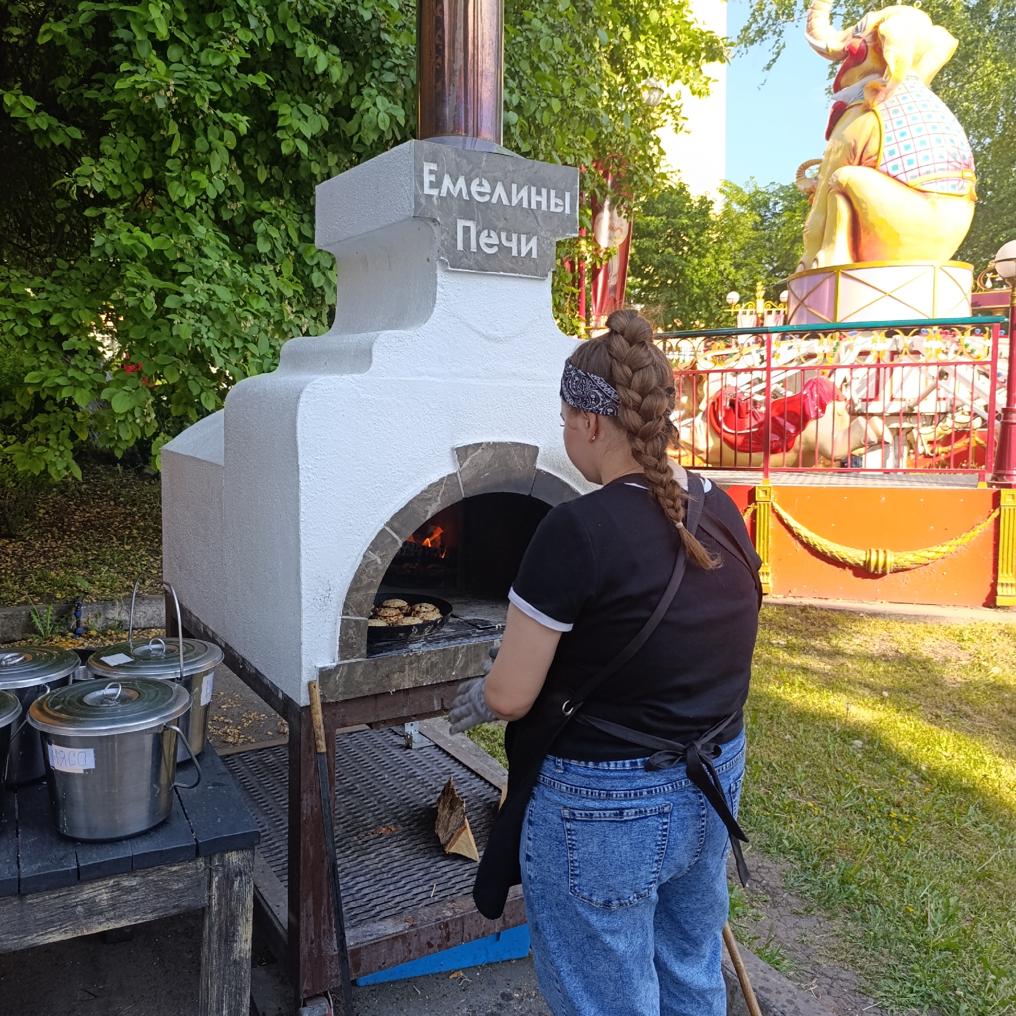 A medium shot captures a woman, likely a cook, tending to a traditional Russian oven, known as "Емелины Печи" ("Emelya's Stoves"). The oven is white with a stone-arched opening, a copper chimney, and a sign that reads "Емелины Печи". Inside, a pan of food cooks over a wood fire. To the left of the oven are metal containers, one labelled "MЯCO" (Meat). The woman, wearing a black t-shirt with white trim, blue jeans, a black apron, and a bandana, is seen from the back, with her hair braided down her back.
The scene is set outdoors, likely in an amusement park, as indicated by the presence of a carousel featuring a large elephant statue in the background. The location is in Izhevsk, Russia. The area is grassy with trees providing shade, suggesting a park or recreational area.