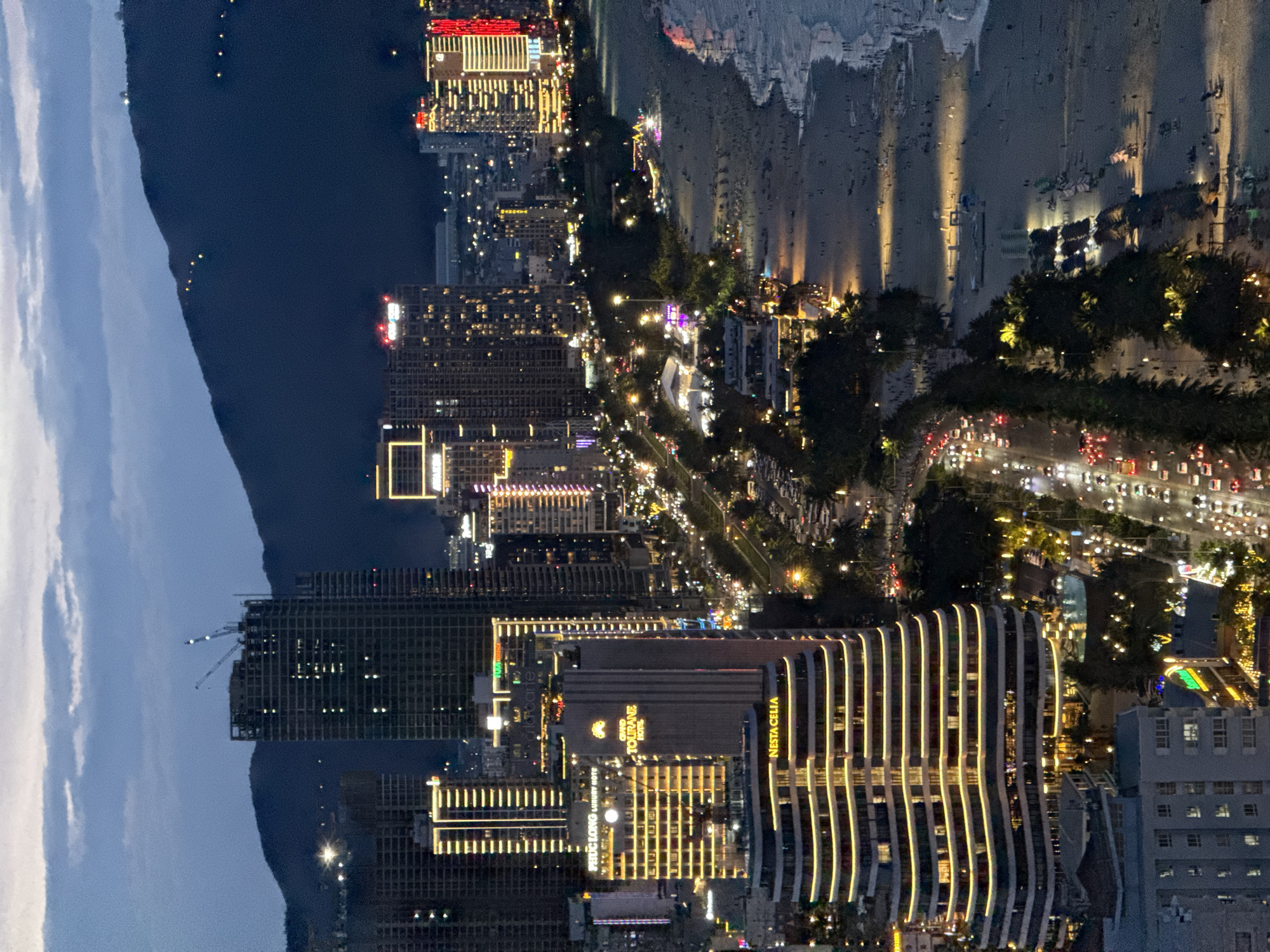 This high-angle shot captures a vibrant coastal cityscape at night or late dusk, overlooking Đà Nẵng, Vietnam. The scene is dominated by a bustling urban landscape flanked by the sea and mountains.
In the foreground and mid-ground, a dense collection of buildings illuminates the night. Many are modern high-rises, their windows and facades glowing with warm light, creating intricate patterns. Prominently visible are several hotels, including "GRAND TOURANE HOTEL," "NESTA CELIA," and "PHÚ LONG LUXURY HOTEL." Another building features the partial text "La Carte Hotel." To the far left, a structure under construction shows cranes against the dark sky.
A wide, busy road runs parallel to the coastline, acting as a bright artery through the city. It is filled with the red taillights and white headlights of numerous vehicles, indicating significant traffic. This road is lined with abundant tropical trees, which are also illuminated, adding pockets of green light to the urban glow.
To the right of the road, a sandy beach stretches along the coast, illuminated by numerous spotlights that cast long, distinct shadows on the sand. The breaking waves of the sea are visible, creating a dynamic edge to the land. The ocean itself is dark, with a few distant lights suggesting other vessels or distant shores.
In the background, a dark silhouette of mountains or hills rises against a sky transitioning from twilight blue to deep night, adorned with scattered clouds. The overall atmosphere is one of a lively, modern city nestled between natural beauty, active well into the evening.