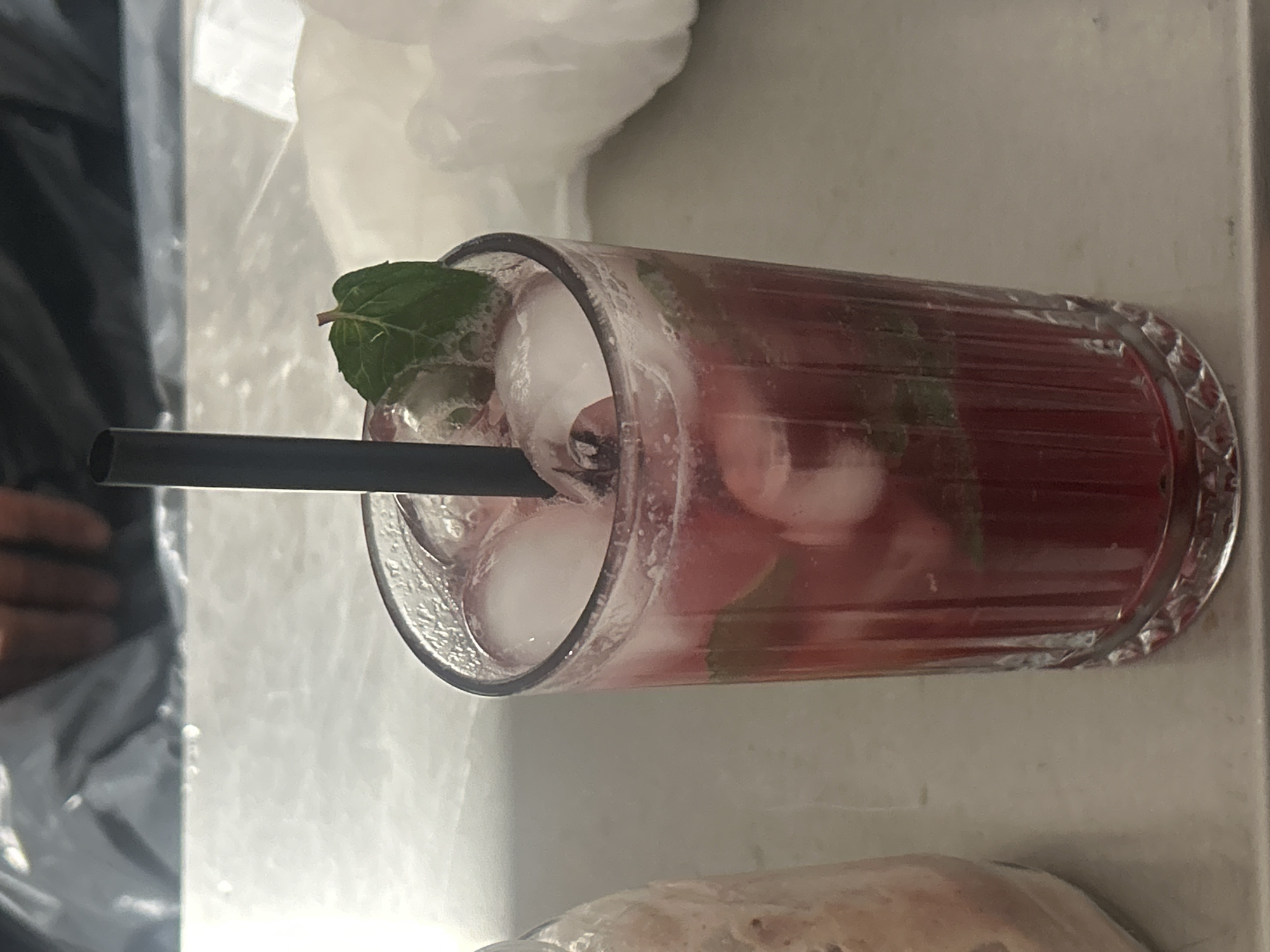 The image shows a cocktail in a glass with ice, a black straw, and a mint leaf garnish. The drink is red. The glass is lying on its side on a table. There is food, a clear plastic bag, and a person's hand visible in the background. Based on prior image analysis, this image was taken at the Flora Bar & Grill in Burgas, Bulgaria.
