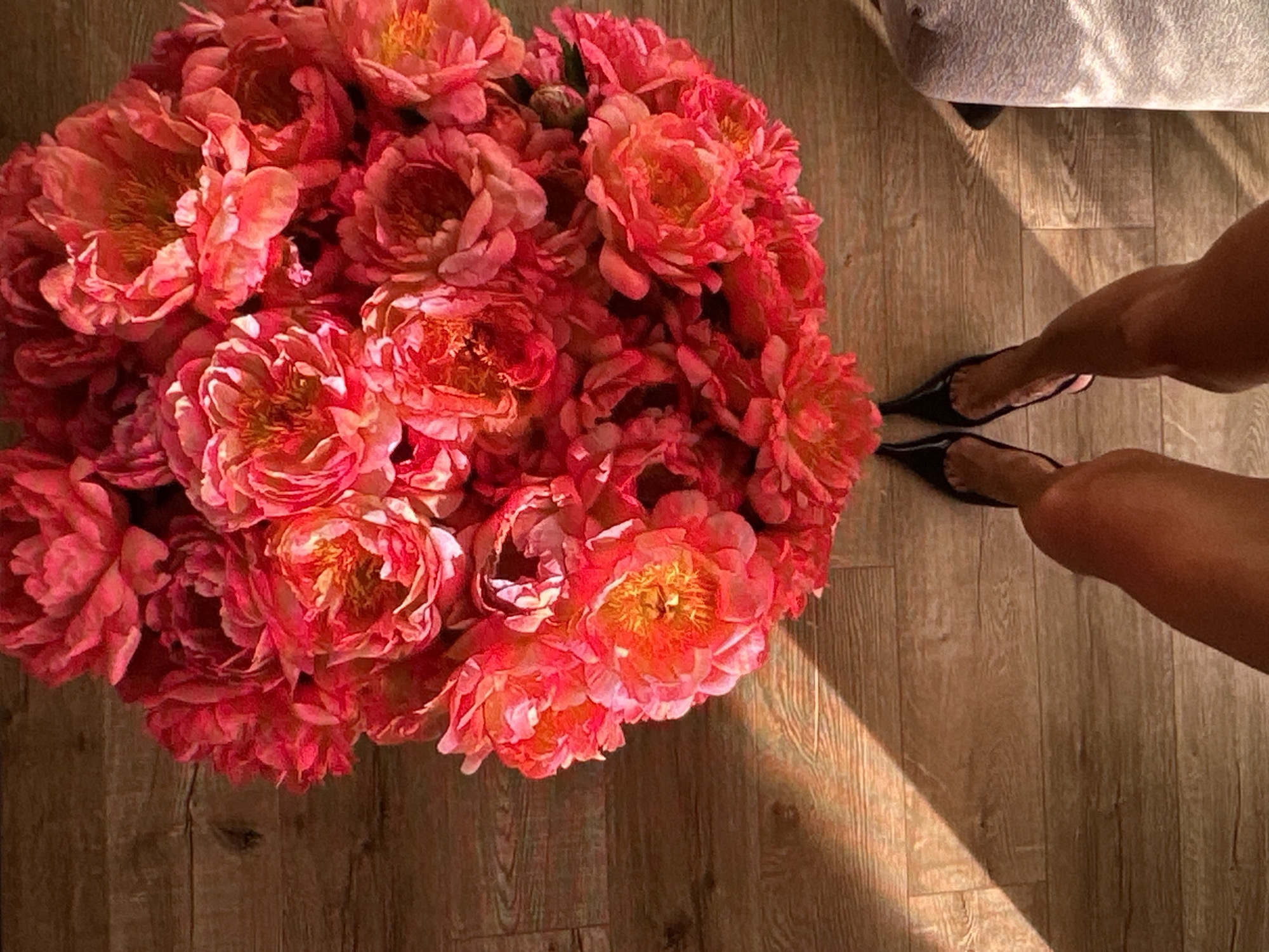 A top-down shot depicts a large bouquet of pink and peach-colored peonies resting on a light-colored wooden floor. In the upper right corner of the frame, a person's legs are visible, clad in black, pointed-toe flat shoes. The scene is illuminated by a bright beam of sunlight. The presence of the patterned flooring and the style of the floral arrangement suggest a domestic interior. The information given is insufficient to determine the specific location in Yerevan, Armenia.