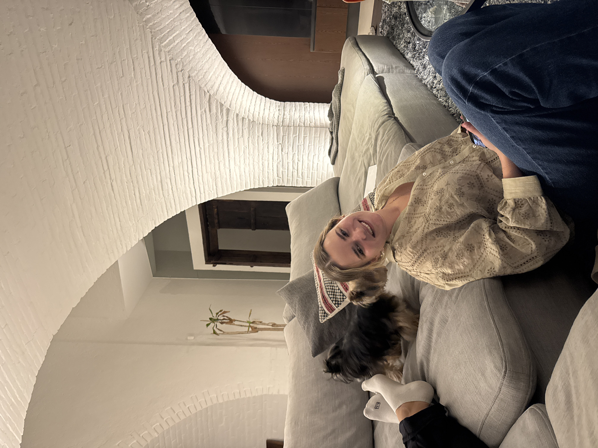 A woman is sitting on a light gray sofa with a small dog, possibly a Yorkshire Terrier, resting on the sofa next to her. The woman is wearing a light-colored patterned blouse. A person wearing blue jeans is also partially visible next to the woman. Another person's feet, clad in white socks and black pants, are visible near the dog. The setting appears to be indoors, possibly in a living room with a white brick archway as an architectural feature. A potted plant can be seen in the background. The location is not evident from the provided image.