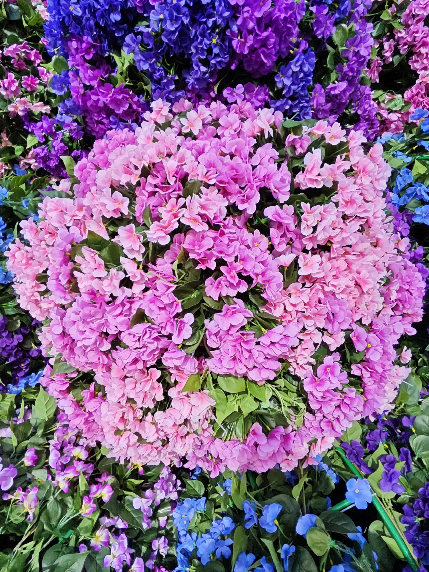 This media file is a close-up, overhead shot of an arrangement of artificial flowers. The subjects are primarily the flowers themselves, displayed in a dense, textured arrangement. The flowers are in various shades of pink, purple, and blue, with green foliage interspersed throughout. A large cluster of bright pink flowers dominates the center of the frame, surrounded by smaller clusters of lighter pink, deep purple, and bright blue flowers. The overall impression is one of vibrant, artificial floral beauty, possibly part of a larger decoration or display. There are no people, text, or specific environmental cues such as time of day or weather visible. The location context of Moscow, Russia, is external information and not discernible from the image itself. The activity taking place is a static display of flowers.