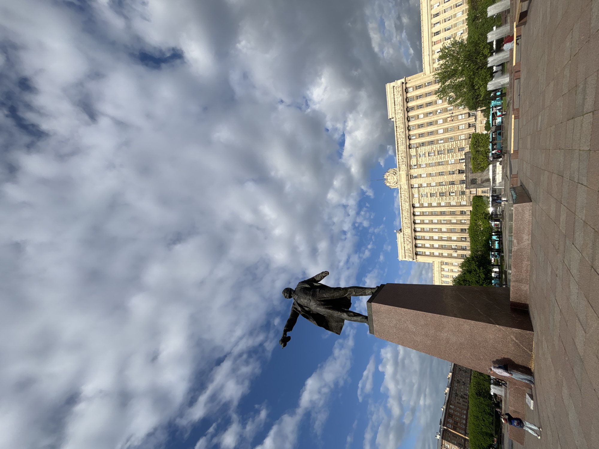 The image depicts a statue of Lenin in Saint Petersburg, Russia. The statue stands on a tall, rectangular pedestal. The background includes a large, ornate building and a sky filled with clouds. The statue is oriented to the left of the scene. The image is taken from a low angle, looking up at the statue and the building. Some people are visible at the bottom right, on the plaza. The plaza is paved with red brick.