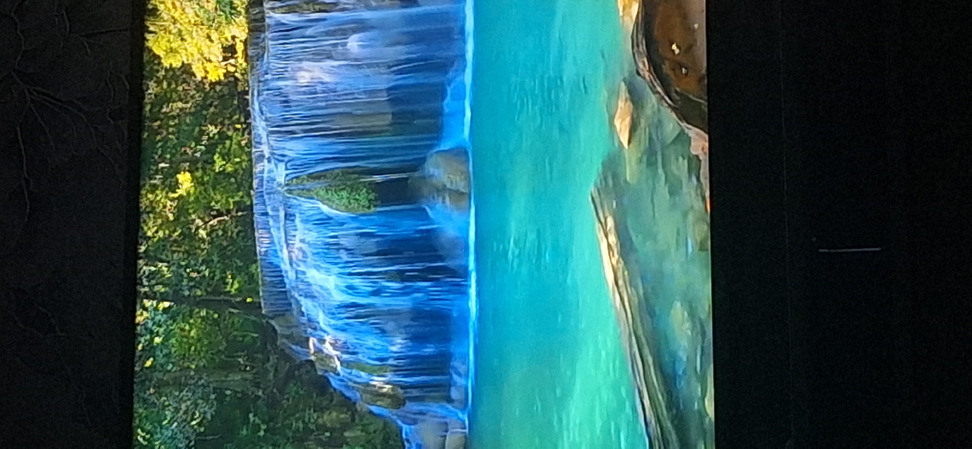 The image displays a picturesque waterfall cascading into a vibrant turquoise pool of water. Lush green foliage and trees form a dense backdrop to the waterfall, suggesting a natural, jungle-like environment. Submerged rocks and a large log are visible in the foreground of the clear water. The top and bottom portions of the image are black, indicating it might be a photograph of a screen or a framed picture. There are no people or animals visible in the scene. The location of this scene is not in Bila Tserkva, Ukraine.