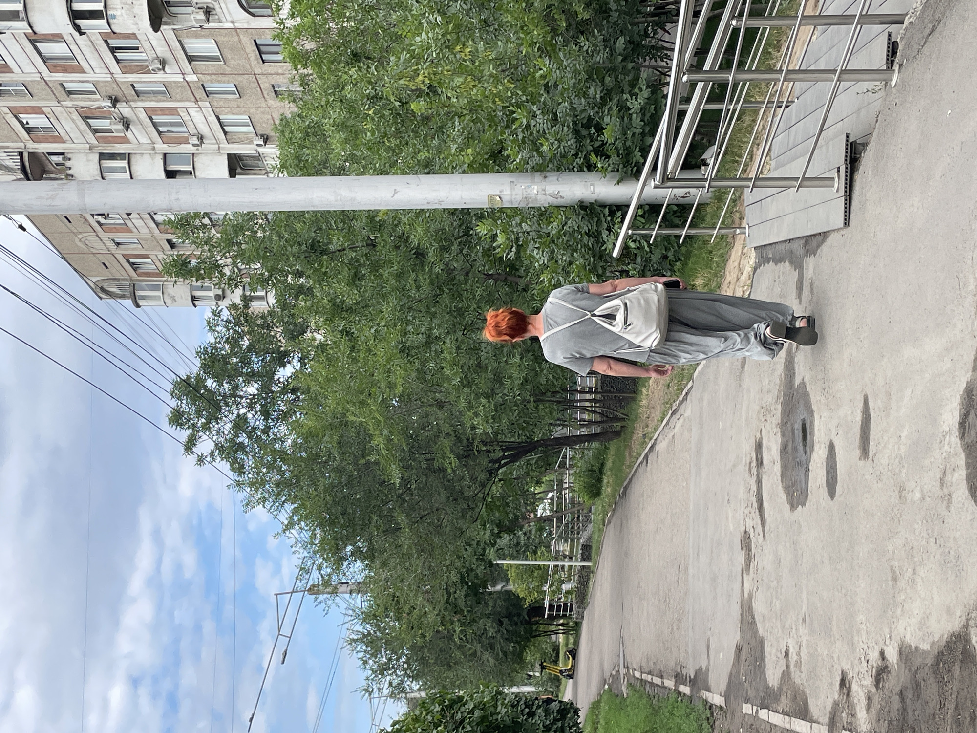 A woman with bright orange hair walks away from the camera on a paved path. She is wearing a loose-fitting gray outfit and carrying a white tote bag over her shoulder. To her left, a yellow electric scooter is parked near some trees. In the background, a multi-story apartment building with numerous windows and air conditioning units stands behind a dense line of green trees. Overhead, power lines crisscross the sky, which is partly cloudy. The scene appears to be in an urban park or residential area.