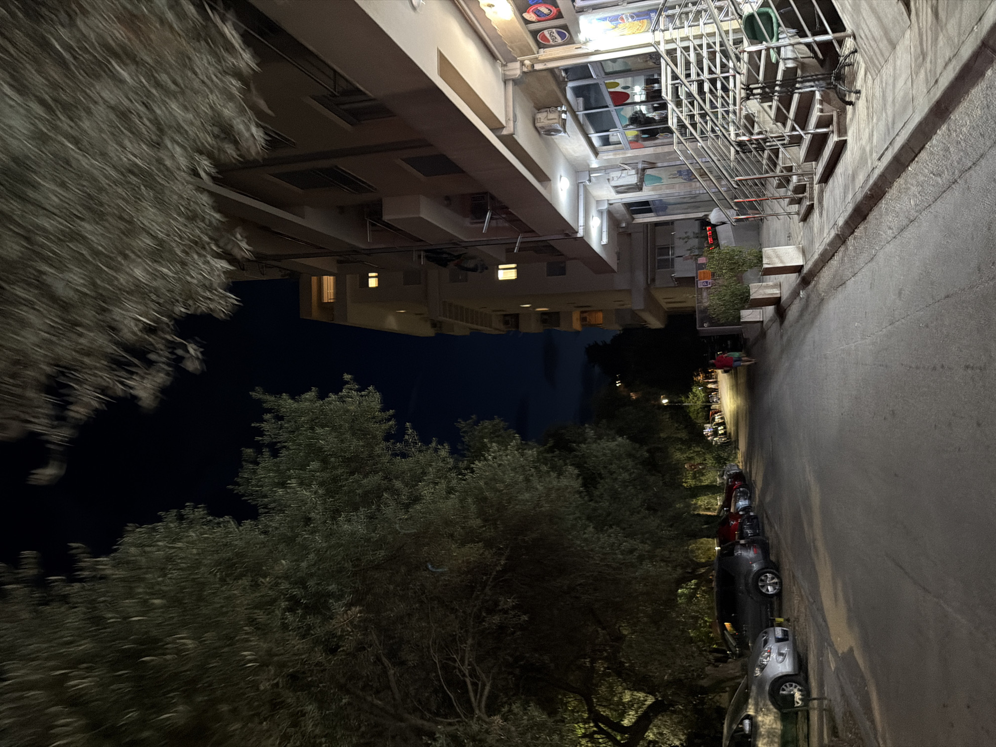 This media file captures a street scene at night in Budva, Montenegro. The image is oriented sideways, with the street running vertically from the bottom to the top of the frame.
The primary subjects include a row of parked cars along the right side of the street, illuminated by ambient light and possibly streetlights. Several cars are visible, some appearing to be sedans or hatchbacks. The left side of the street is bordered by trees, their foliage dark against the night sky, with some lights illuminating the lower branches.
Behind the trees and extending along the right side of the street is a building with multiple floors. The building's facade is light-colored, and some windows are illuminated from within, suggesting occupancy. On the upper floors, there is a section with a metal grate structure and various signs and objects, possibly indicating shops or businesses. Some of these signs feature logos, including one that appears to be for Pepsi.
The scene suggests a quiet evening in a residential or mixed-use area. The time of day is clearly night due to the darkness of the sky and the artificial lighting. The weather appears calm, with no indication of rain or wind. The location cues of Budva, Montenegro, place this scene in a coastal European city.
Notable details include the motion blur in the trees on the left, suggesting a slight breeze or movement within the frame. The overall mood is subdued, typical of a nighttime street view. There is no overt emotional expression or activity depicted, focusing more on the static elements of the urban environment.
Visible text includes the Pepsi logo and potentially other brand names or advertisements on the building's facade, though these are not clearly legible.