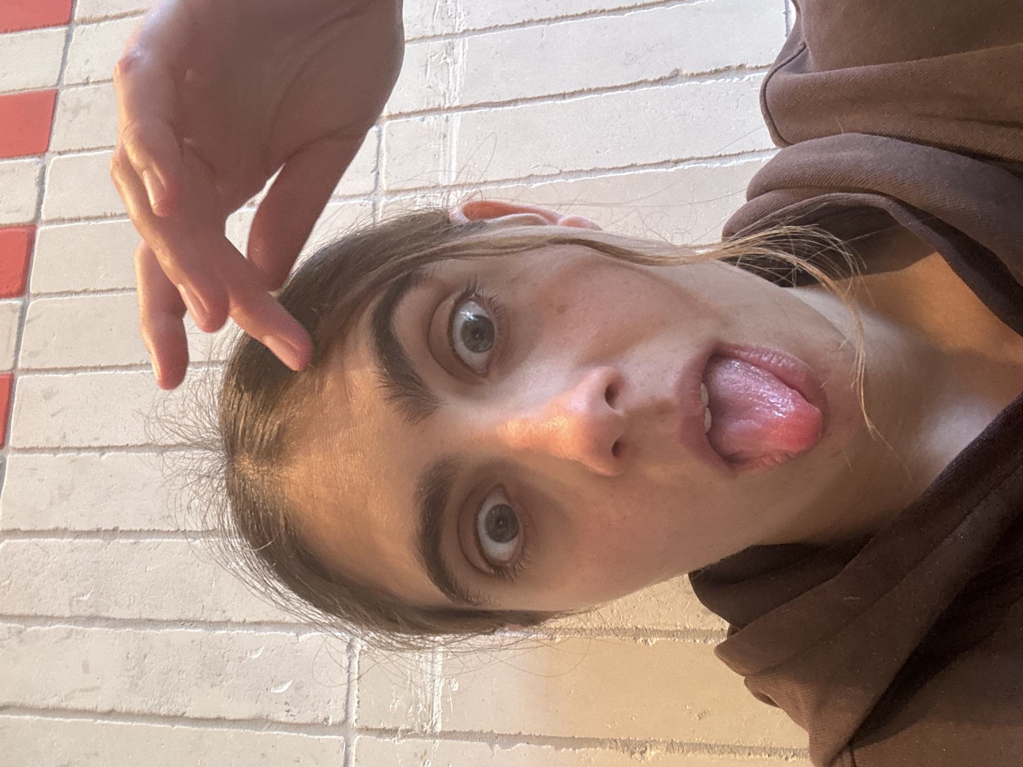 A person with fair skin and dark eyebrows is sticking out their tongue at the camera. They are wearing a brown hoodie. A hand is partially visible above their head, with fingers pointing towards their forehead. The background consists of white and red tiles. The location is not identifiable as Tel-Aviv, Israel based solely on the image.