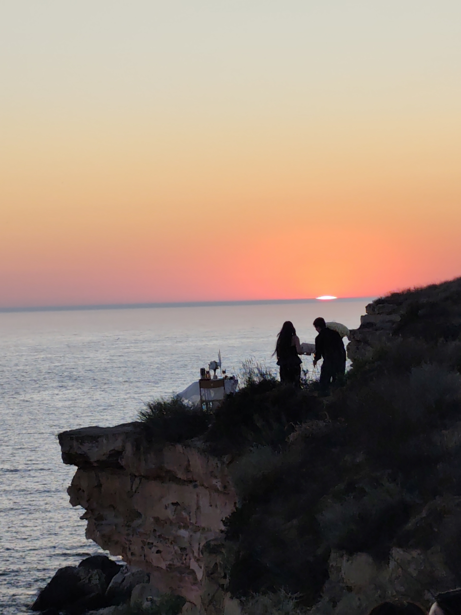 The image depicts a sunset scene over the Caspian Sea, likely near the city of Aktau, Kazakhstan. The sky is a gradient of soft yellows, oranges, and pinks, with the sun a bright, glowing disc just touching the horizon. In the foreground, silhouetted figures of a couple stand on a cliff overlooking the sea. The man, wearing dark clothing, appears to be interacting with the woman, who is also dressed in dark attire and has long hair. They are positioned on a rocky, vegetated cliff face. To the left, a small structure with some equipment, possibly a navigational aid or a small observation post, is visible on the cliff edge. The sea is calm and reflects the colors of the sky. The overall impression is one of a serene, natural landscape at dusk.