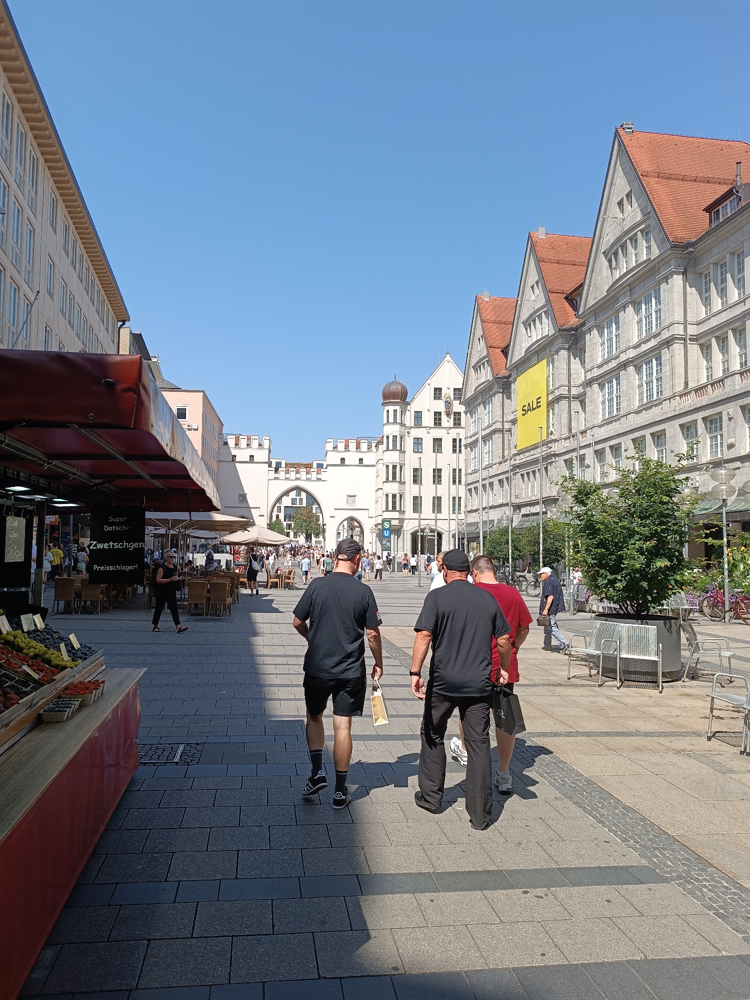 The image depicts a street scene in Munich, Germany, likely a shopping area given the presence of market stalls and shops. In the foreground, three men are walking away from the camera on a paved pedestrian walkway. The man on the left wears a black t-shirt and black shorts. The man in the middle wears a red t-shirt and black pants. The man on the right wears a black t-shirt and grey pants. To the left, a market stall with a red awning displays various fruits. Beyond the stall, outdoor seating for a cafe or restaurant is visible with patrons seated at tables.
In the midground, more pedestrians are walking, and a prominent archway is visible in the distance, suggesting it is a significant landmark, possibly part of a historical structure or entrance to a square. The architecture on the right side of the street features multi-story buildings with stone facades and red tile roofs, typical of older European city architecture. A large yellow banner with the word "SALE" is displayed on one of the buildings. Trees in planters are placed along the walkway, and bicycles are parked on the side. The sky is clear and blue, indicating a sunny day. The overall atmosphere appears to be one of a busy, commercial city center on a pleasant day. The specific location is likely near the Marienplatz or Sendlinger Tor, based on the architectural style and the presence of the archway.