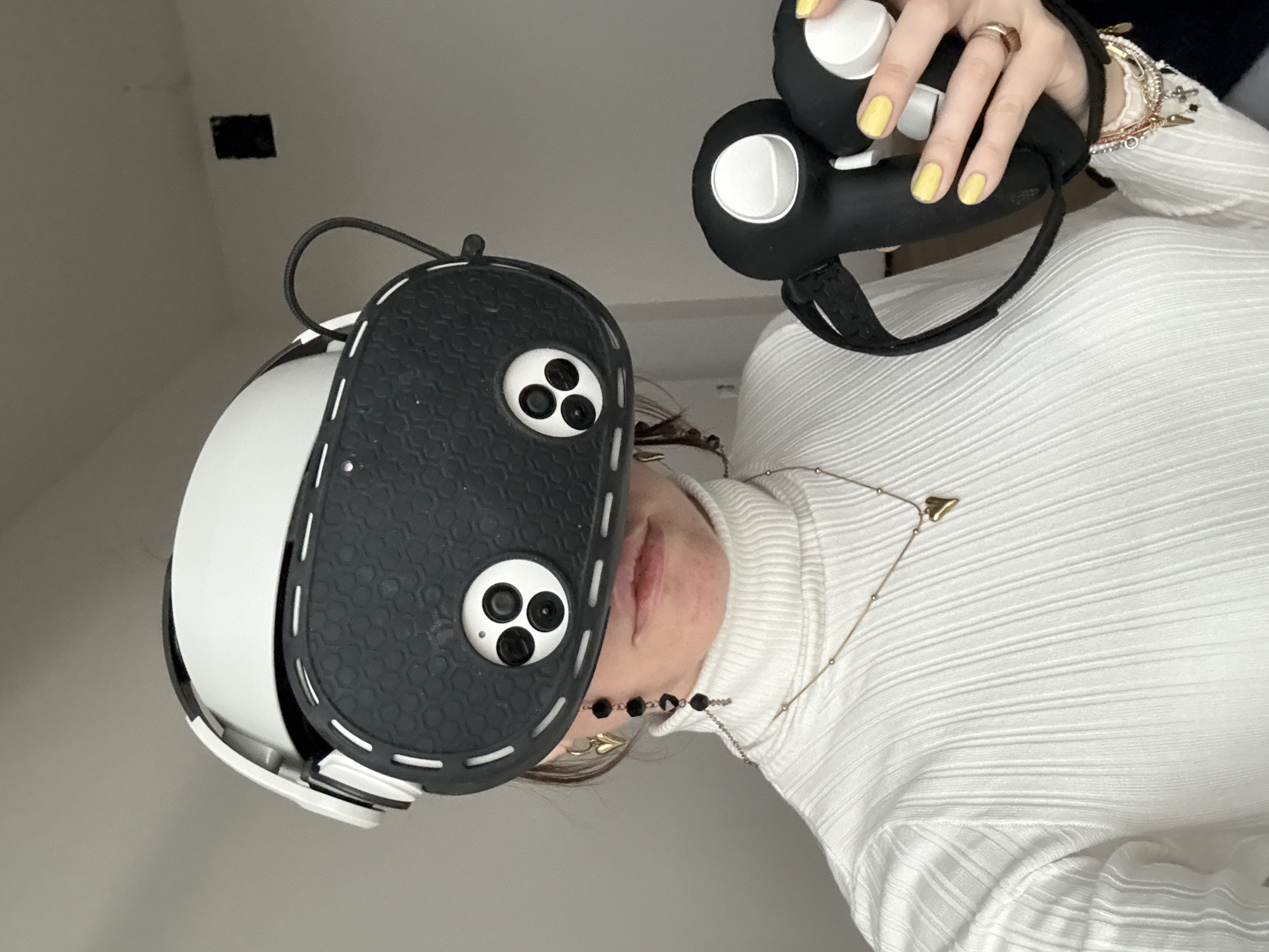 A person is holding a VR headset and controllers. The headset is white with a black faceplate. The controllers are black with white buttons. The person is wearing a white turtleneck sweater and has yellow nail polish. The background is a plain white wall with a small square cutout. The location is not evident from the image.