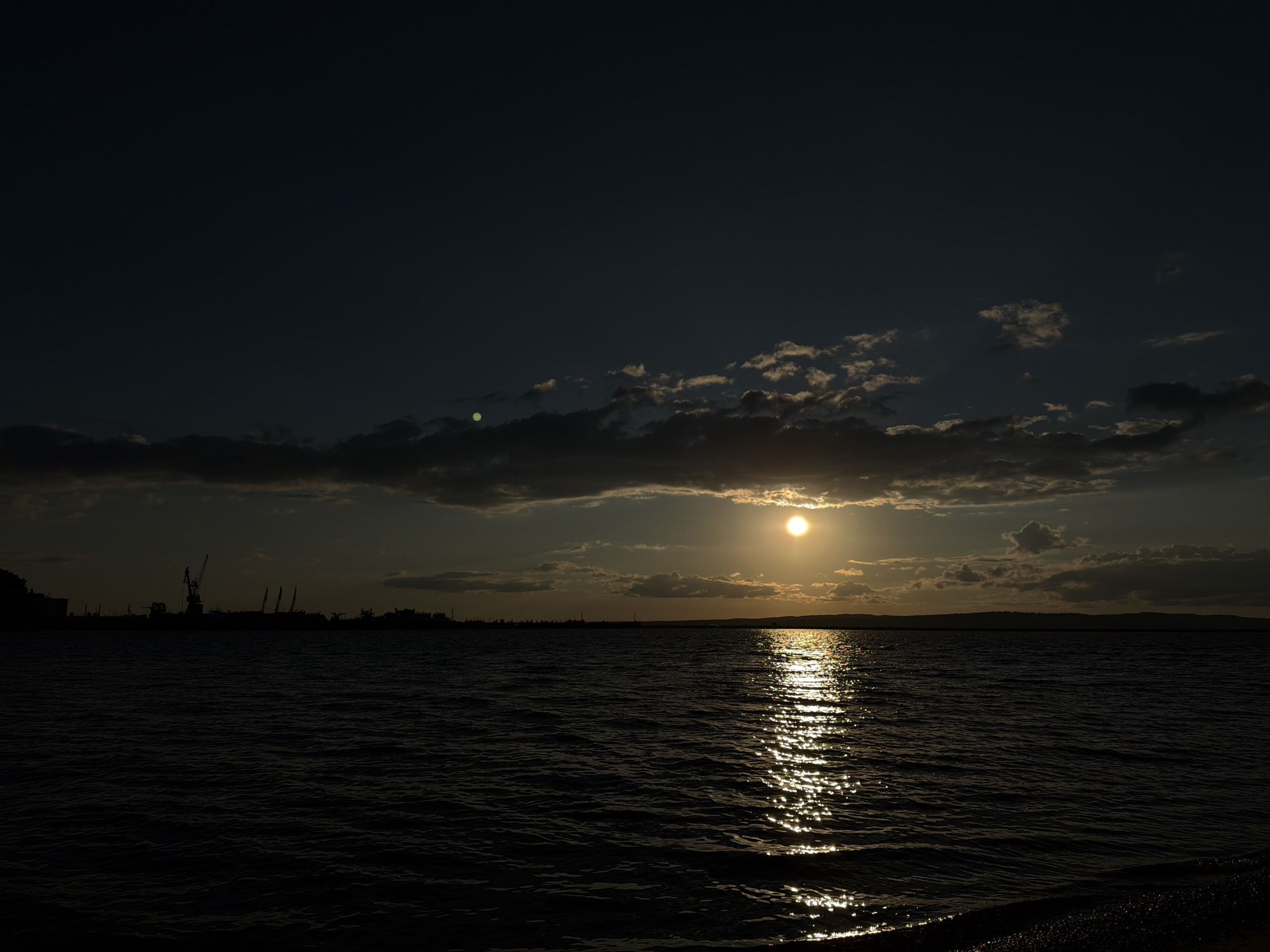 The image depicts a sunset over the Kama River in Chaikovsky, Russia. The sun, a bright orb, is partially obscured by clouds on the horizon, casting a shimmering reflection on the water. Silhouettes of industrial cranes and buildings are visible on the left side of the image, suggesting a port or industrial area. The sky is a deep blue with scattered, wispy clouds illuminated by the setting sun. The water is dark and choppy, with small waves. The foreground shows a dark, pebbled shoreline. There are no people or animals visible in the scene.