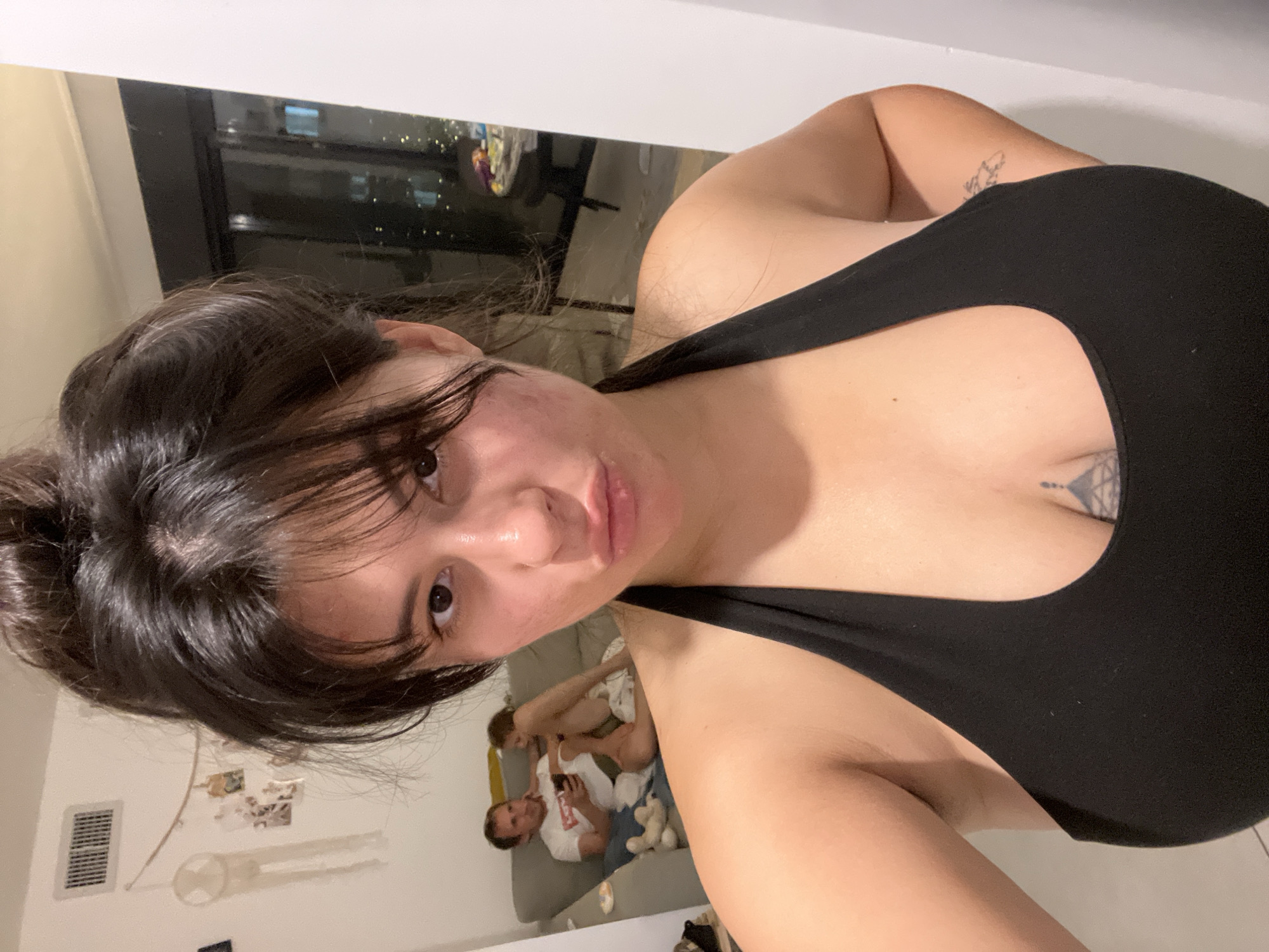 A selfie taken indoors shows a woman in the foreground with her hair in a messy bun, pouting her lips. She is wearing a black halter top. In the background, a man and a child are sitting on a couch. The man is wearing a white t-shirt and looking at his phone. The child is partially visible, facing away from the camera. The room appears to be a living area, with a vent on the wall and a window in the background showing a city skyline at night, suggesting the location is Kiryat Bialik, Israel. There are also some decorative items on the wall and a table with items on it visible through the window.