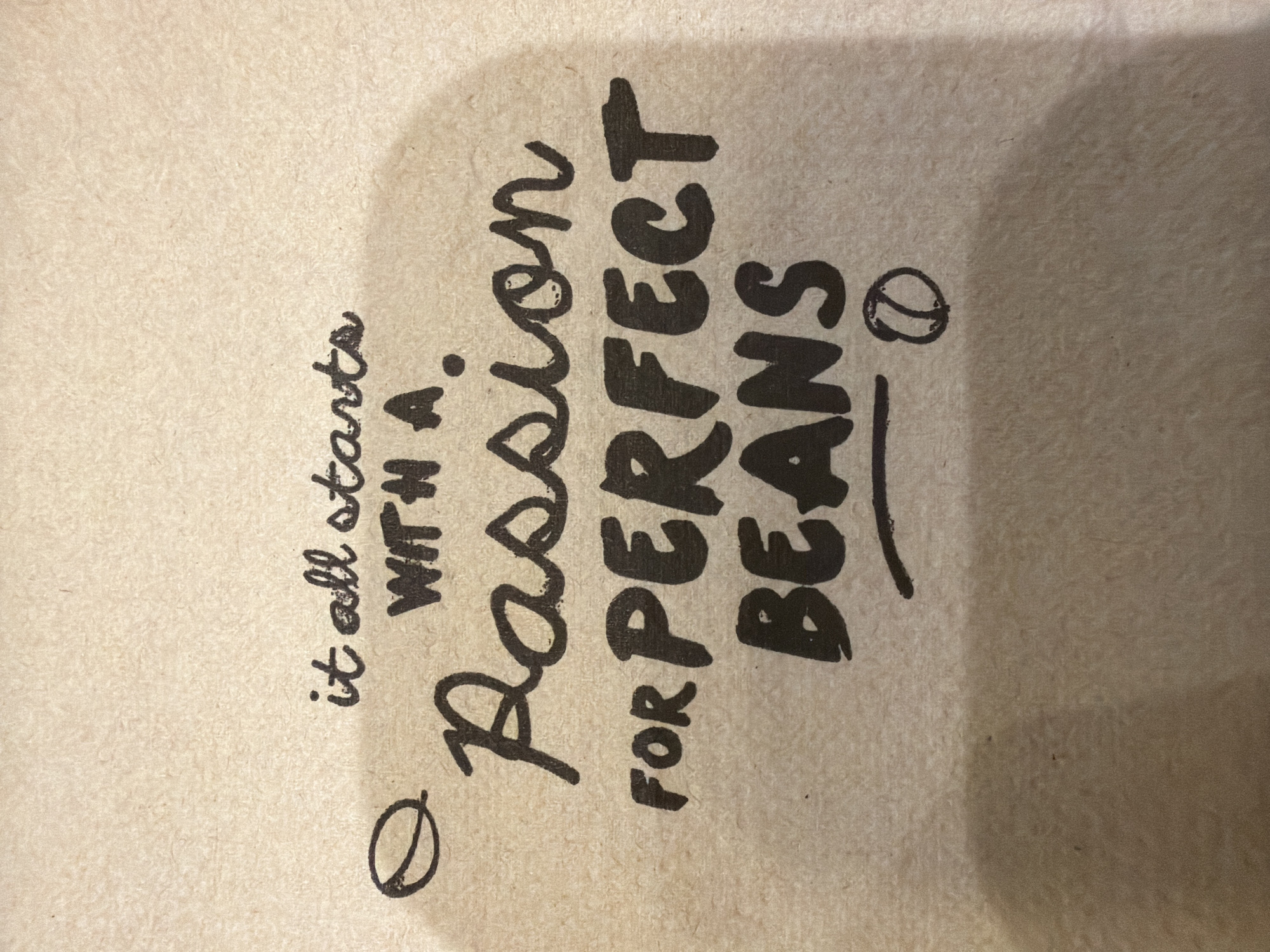 The image shows a close-up of a beige, textured surface with black text. The text reads "it all starts WITH A Passion FOR PERFECT BEANS". The letters are in a mix of cursive and print styles, and some words are larger than others. There is an illustration of a coffee bean to the left and right of the text. The location in the city of Mole Valley, United Kingdom is not apparent from the image.