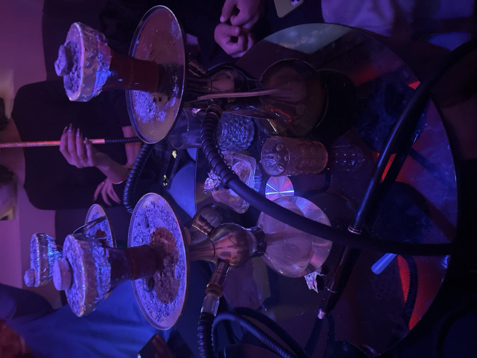 The image depicts an indoor scene in the city of Paris, France, characterized by dim ambient lighting dominated by blue and red tones. A dark, reflective table serves as the primary surface, upon which two active hookahs are centrally positioned. The hookah on the left features a brass-toned stem and a clear glass base, with its bowl covered in aluminum foil containing burnt shisha tobacco and a glowing charcoal piece. A black hose is connected to it. The hookah on the right, also with a brass-toned stem, has an amber-tinted glass base, and its foil-covered bowl contains similar shisha material and charcoal.
Three individuals are partially visible. In the mid-right, a person with dark hair and dark-painted fingernails is holding a hookah hose. In the background left, an individual wearing blue attire is partially observed. The hands and forearms of a third person are visible in the lower right quadrant.
Additional objects on the table include multiple patterned drinking glasses (one clear, one reddish-orange), a small clear square container, a black pen-like instrument, and a small reflective disc. The arrangement of objects and people suggests a social gathering in a lounge or shisha bar setting.