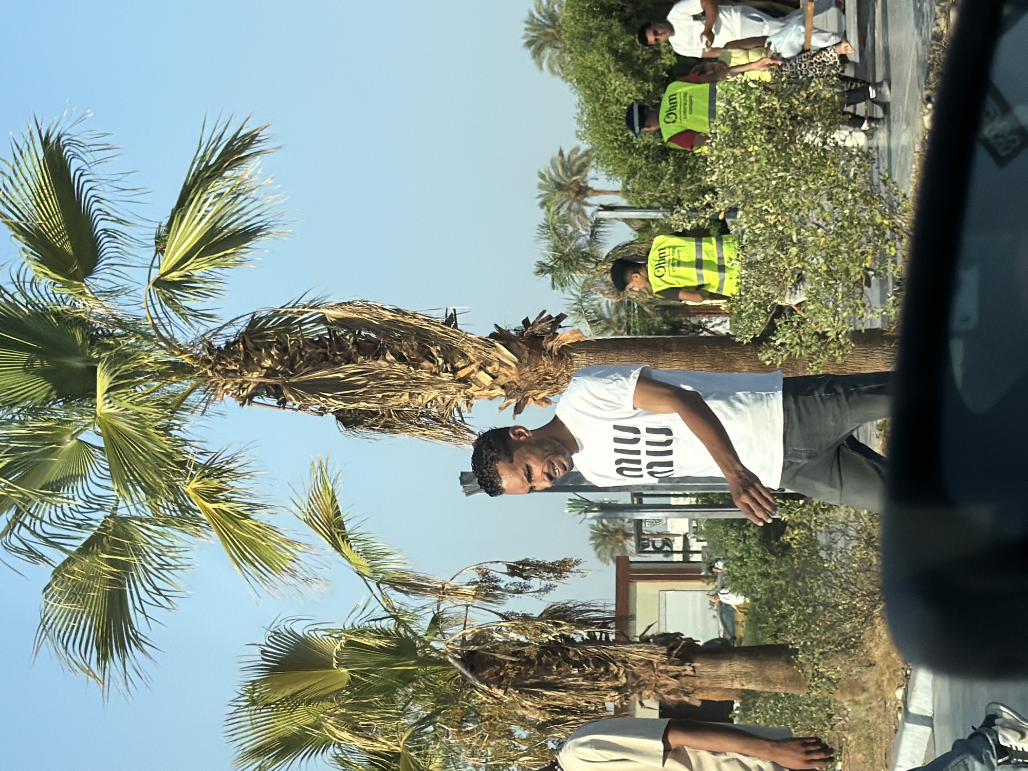 In Sharm El Sheikh, Egypt, an outdoor scene unfolds beneath a clear blue sky, dominated by the presence of lush palm trees with their distinctive fronds. In the foreground, a man in a white t-shirt with a graphic design and dark pants walks towards the right, his gaze directed away from the camera. To his left, another person, partially visible, stands facing left.
In the mid-ground, a man wearing a bright yellow safety vest with the logo "Ohm" is visible, as is another individual in a similar vest. Further to the right, a man in a white t-shirt and sunglasses holds a mobile phone, and a woman in a patterned skirt walks behind him. The environment is characterized by greenery, including bushes and more distant palm trees, suggesting a public outdoor space or a resort area. The overall impression is one of a sunny day in a tropical or sub-tropical urban setting.