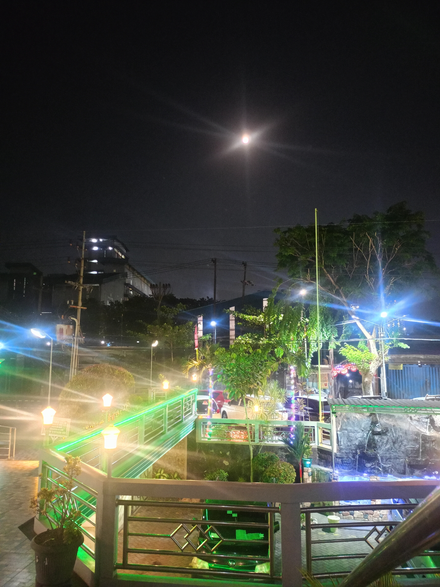 The image depicts an outdoor scene at night, likely in the city of Sidorukun, Indonesia. The sky is dark with a bright moon visible in the upper center, emitting a lens flare. In the foreground, a metal railing with a diamond pattern borders a walkway. A potted plant is positioned to the left of the walkway.
A brightly lit green railing lines a sloped walkway or bridge, with a series of lamp posts illuminating the path. These lamps have a hexagonal shape with glowing elements inside. Further back, trees with illuminated foliage are visible, with bright blue lights casting a glow on them and surrounding structures.
In the mid-ground, buildings with illuminated windows suggest commercial or residential use, with power lines running overhead. A man-made waterfall feature is present to the right, with water cascading over rocks into a pool below, illuminated by blue lights. Vehicles, including a car and a three-wheeled vehicle, are parked or driving in the background. The overall atmosphere is festive and illuminated, suggesting a public space or entertainment area.