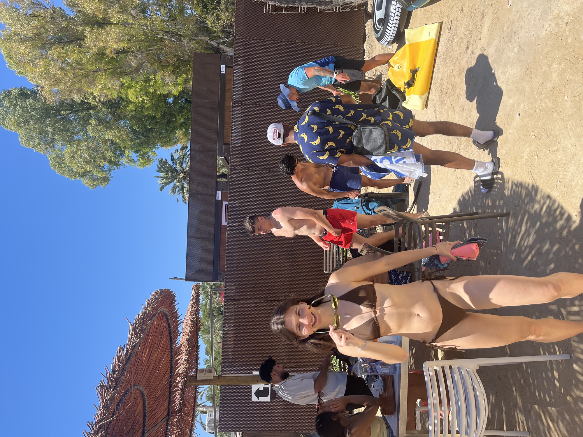 The image shows a beach scene, likely in Teulada, Spain. In the foreground, a woman in a brown bikini stands at a table, holding sunglasses. Behind her, several men are standing in line. One man is shirtless wearing red shorts, another wears blue swim trunks. A third man wears a blue shirt with bananas and white cap. A fourth man is wearing blue shorts and a blue cap and is bending over a yellow object. The area appears to be a beach with a sandy ground. To the left is a thatched umbrella providing shade. A metal structure is behind the men. The sky is clear and blue. Trees are present near the metal structure.