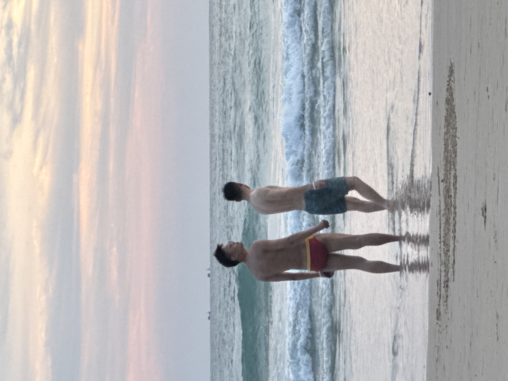 In Karon, Thailand, two shirtless men stand knee-deep in the ocean at the beach. The man on the left, facing away from the camera, wears red swim briefs with a yellow waistband. The man on the right, also facing away, wears dark swim shorts. The ocean has moderate waves and the sky above is filled with pastel-colored clouds, suggesting it is either sunrise or sunset. The beach in the foreground is sandy, with some debris visible. In the distance, two small boats are visible on the horizon.