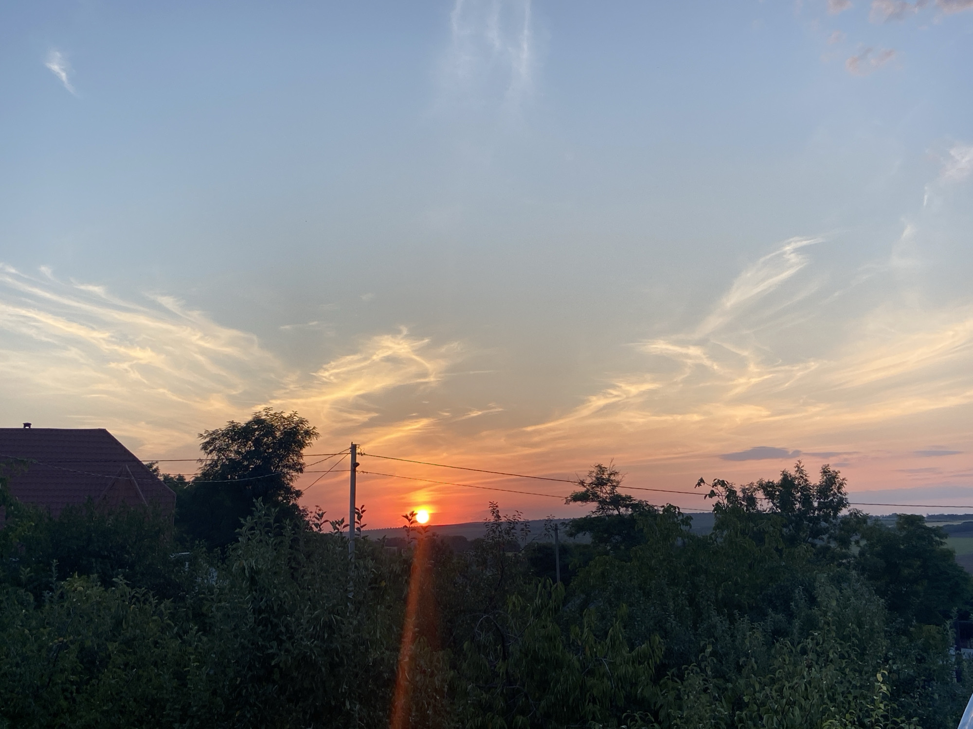 A wide shot captures a serene sunset scene in a rural setting. The sky is a gradient of soft blues and warm oranges, with wispy clouds streaked across the horizon. The sun, a bright orange orb, is positioned just above the tree line, casting a warm glow. The foreground is dominated by lush green trees and foliage, with the red-tiled roof of a house visible on the left side of the frame. A utility pole with wires stands tall amidst the trees, a subtle human element in the natural landscape. The overall impression is one of tranquility and the beauty of nature.
