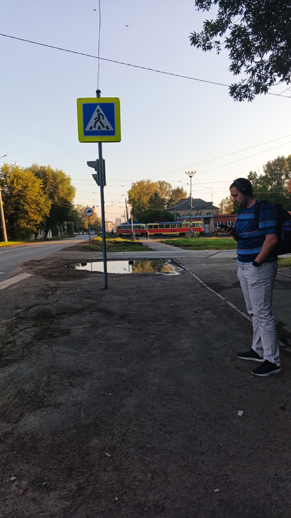 In Izhevsk, Russia, a man with headphones and a backpack is standing on a paved area near a road. He is looking at his phone, with his right hand in his pocket. He is wearing a blue and white striped t-shirt, light-colored pants, and dark shoes. A pedestrian crossing sign is visible on a pole behind him. Across the road, a red and yellow tram is stopped. The environment appears to be urban, with trees and buildings visible in the background. The sky is clear and the light suggests it is either early morning or late evening.